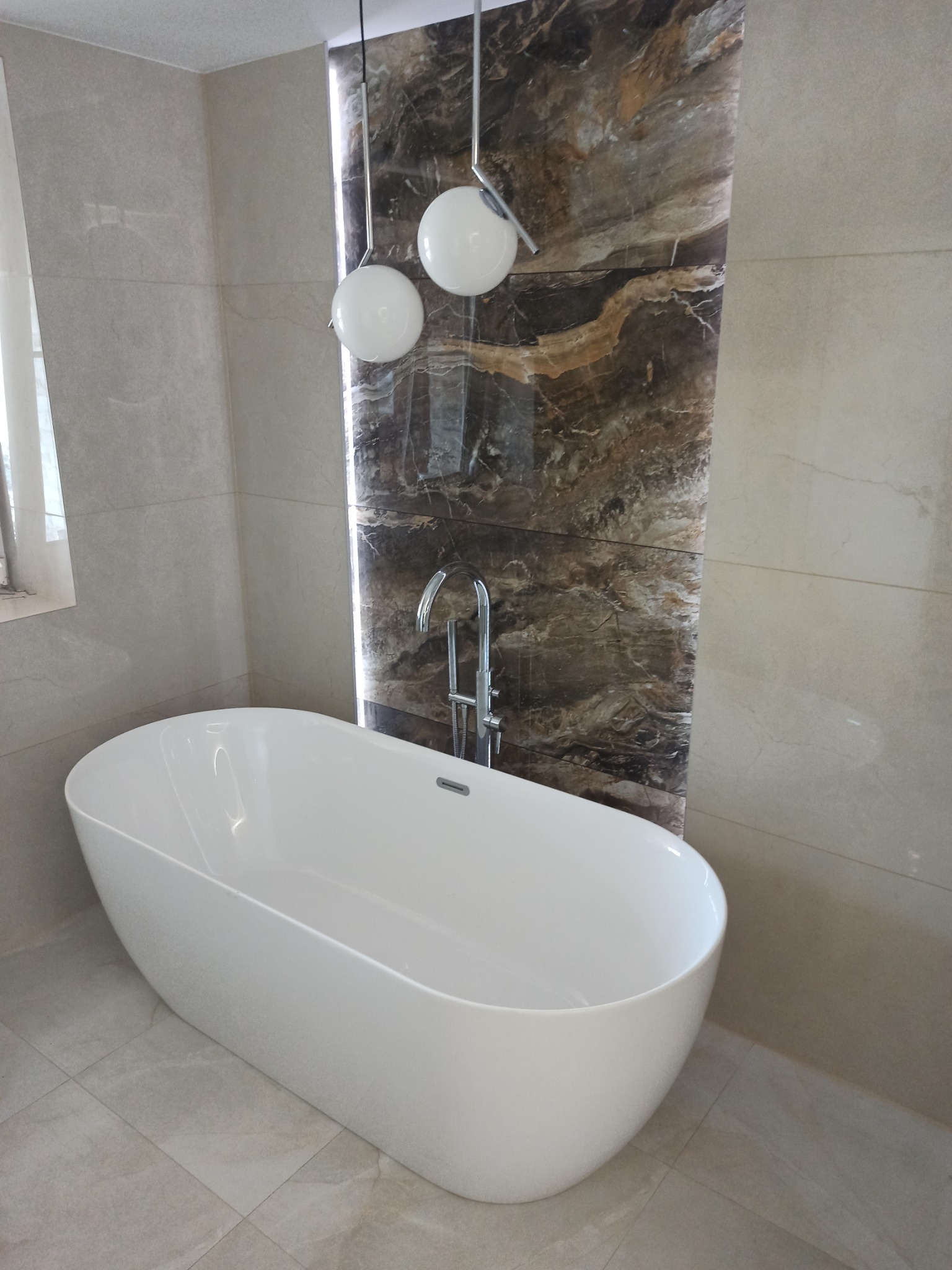 Wolnostojąca wanna w nowoczesnej łazience z beżowymi płytkami i panelem z imitacją marmuru, oświetlona wiszącymi kulistymi lampami.