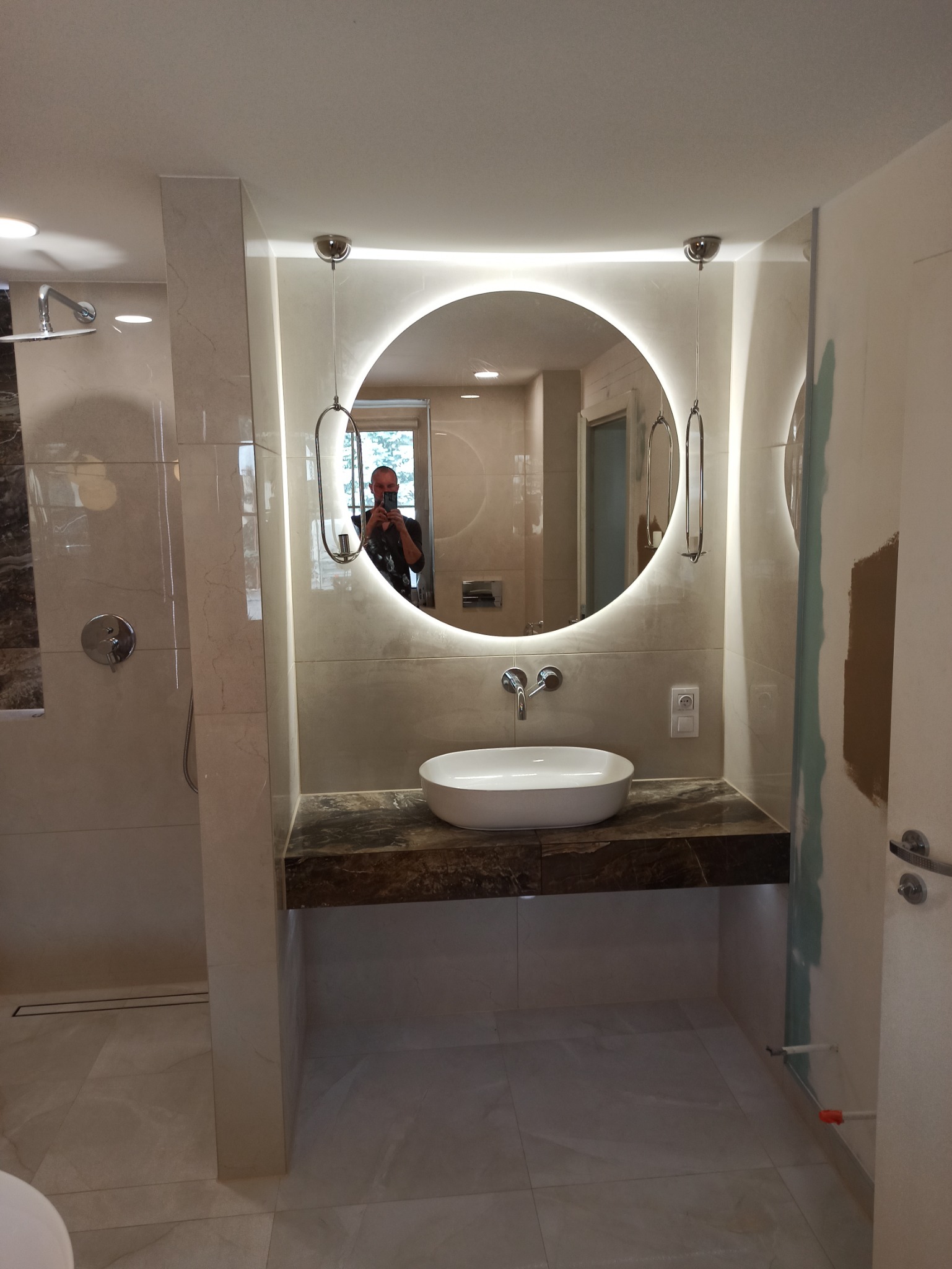 Nowoczesna łazienka w Nakle nad Notecią z okrągłym lustrem z podświetleniem LED, marmurowym blatem i białą umywalką nablatową. Widoczne świeżo położone płytki i fragment ściany przygotowanej...