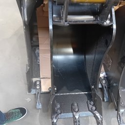 Dom TECH Szymon Sikora - Nowy, czarny, błyszczący osprzęt do koparki, łyżka z zębami, widoczne detale mocowań i śruby, na szarym betonowym podłożu, obok noga w zielonym sportowym bucie.