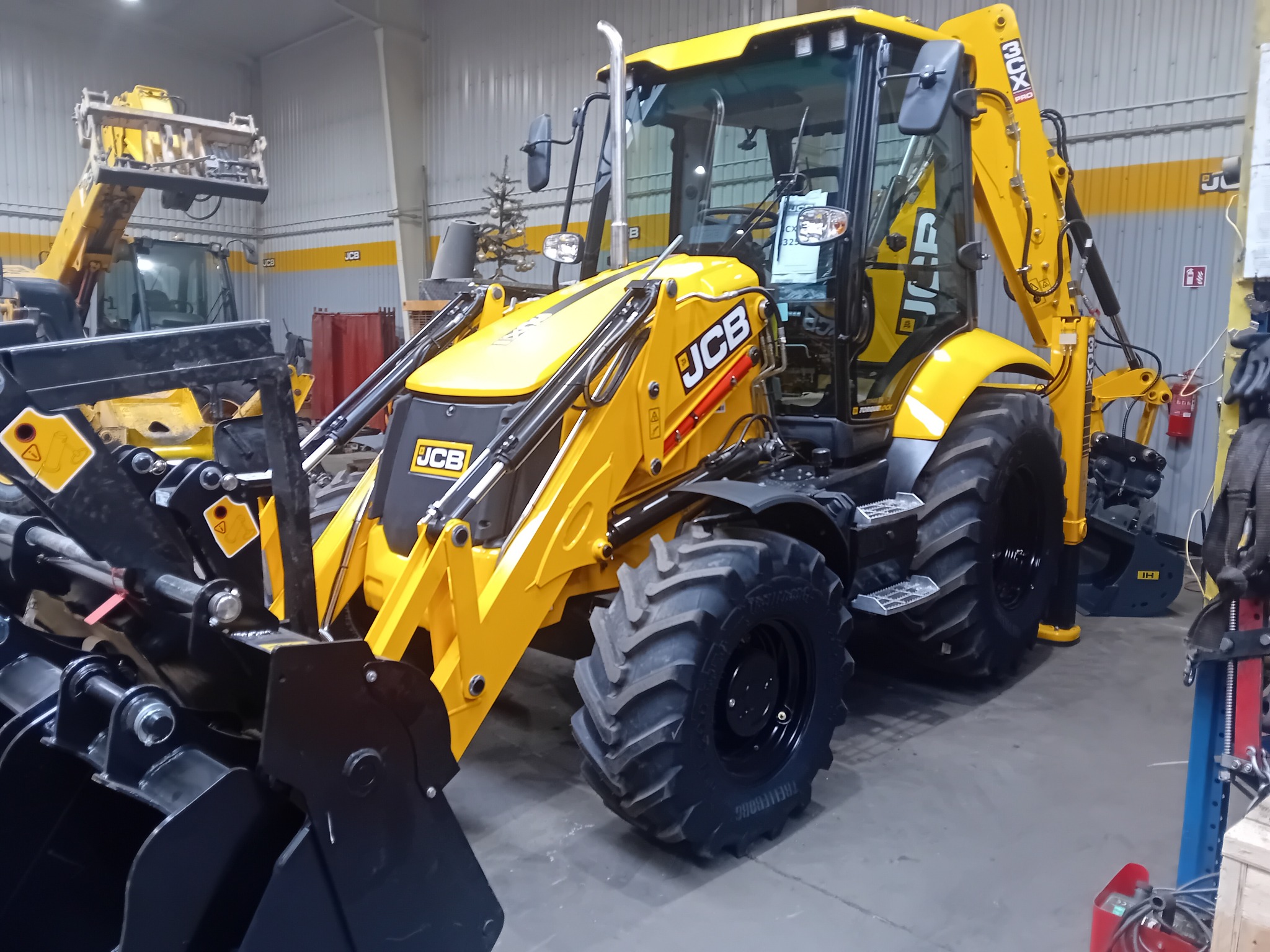 Żółta koparko-ładowarka JCB w hali, widoczny czarny osprzęt z przodu, opony Trelleborg i częściowo inne maszyny w tle.