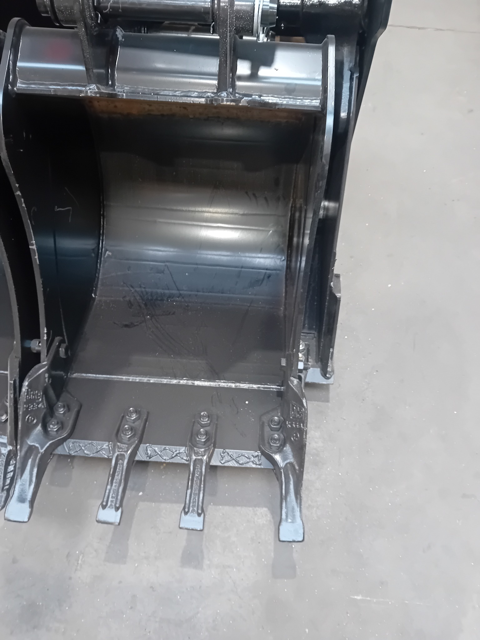 Dwa czarne, metalowe łyżki koparki z zębami, ustawione obok siebie na betonowej podłodze w magazynie.