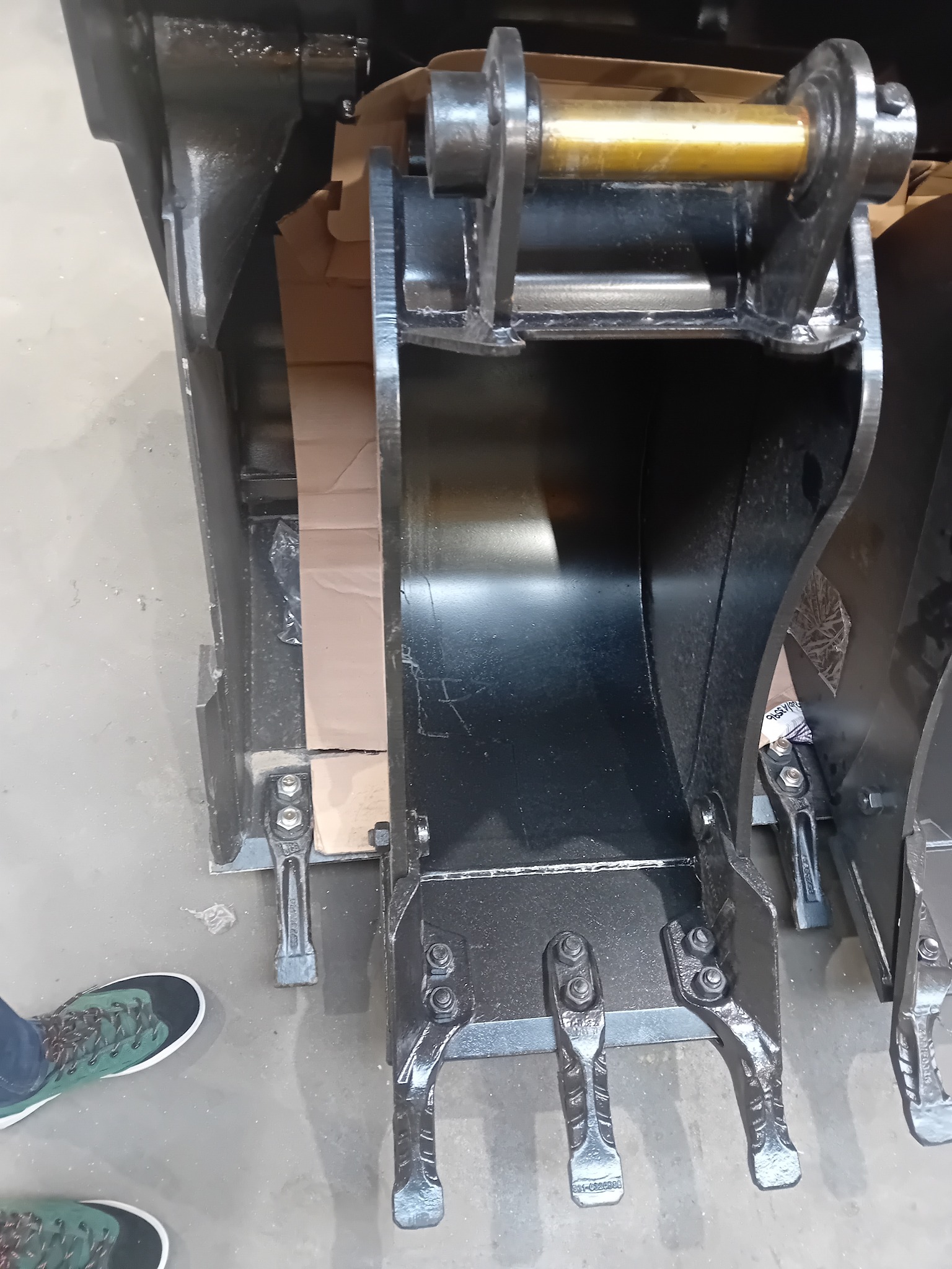 Nowy, czarny, błyszczący osprzęt do koparki, łyżka z zębami, widoczne detale mocowań i śruby, na szarym betonowym podłożu, obok noga w zielonym sportowym bucie.