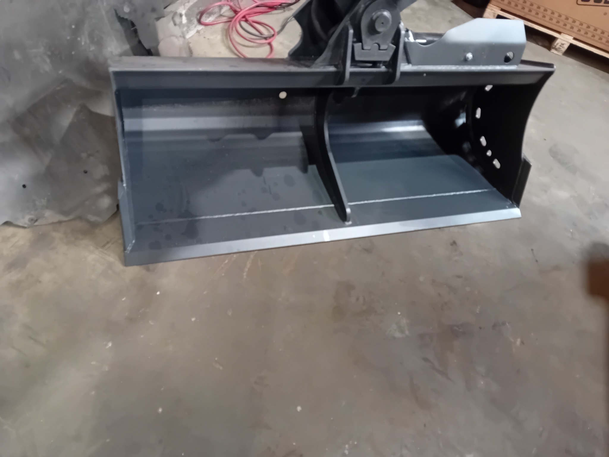 Nowa, szara łyżka do koparki, z widocznymi otworami bocznymi i mocowaniem, leżąca na betonowej posadzce w magazynie.