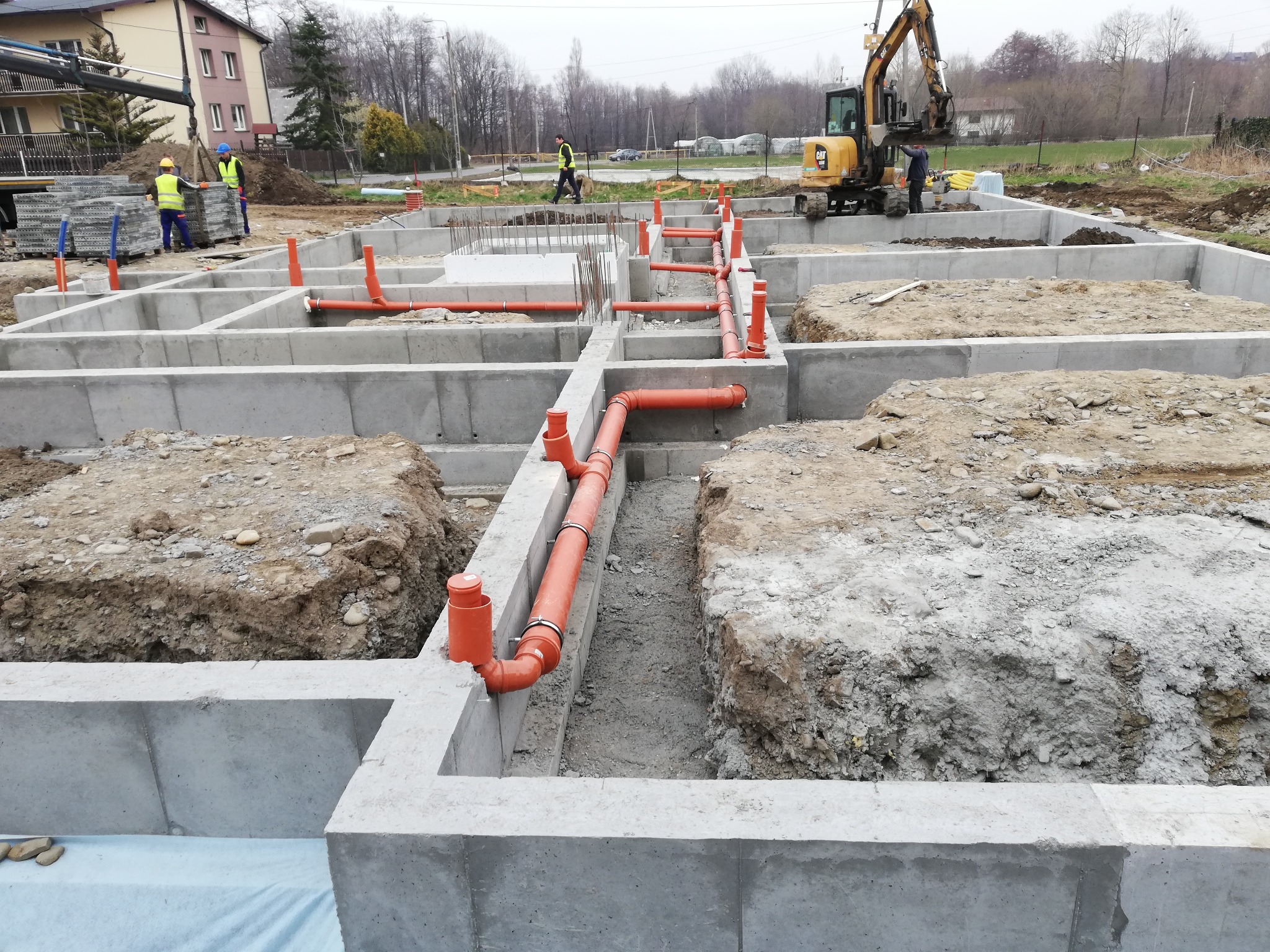 Betonowe fundamenty domu w trakcie budowy z pomarańczowymi rurami kanalizacyjnymi, koparka w tle, pracownicy w odblaskowych kamizelkach przy rozładunku materiałów budowlanych.