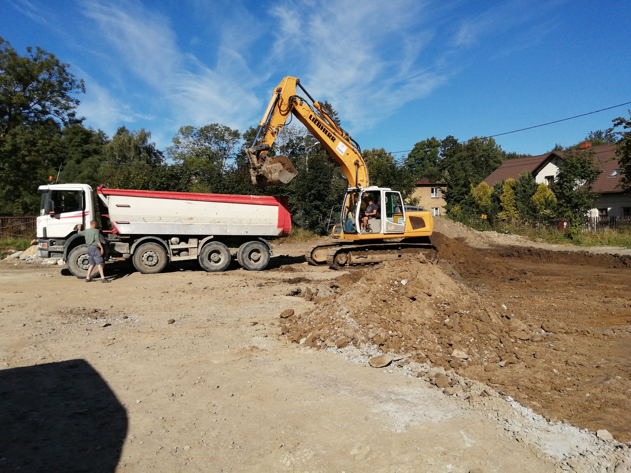 Żółta koparka Liebherr nabiera ziemię i ładuje na białą wywrotkę z czerwoną burtą, na tle błękitnego nieba z chmurami i zieleni drzew oraz zabudowań.