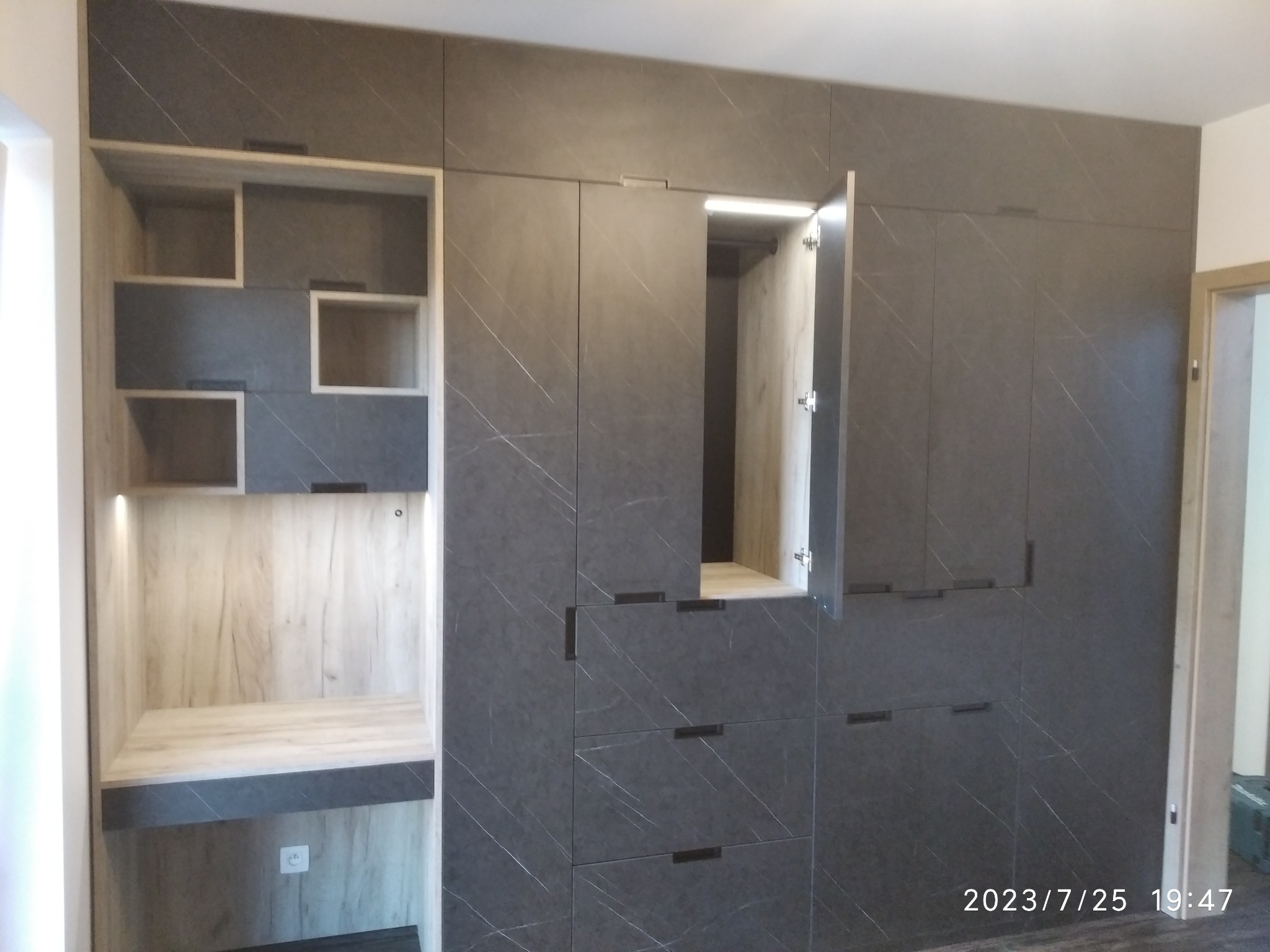 Duża, zabudowana szafa z szarymi frontami imitującymi marmur, z otwartą szafką i półkami oświetlonymi od wewnątrz, na tle ściany i futryny drzwi.