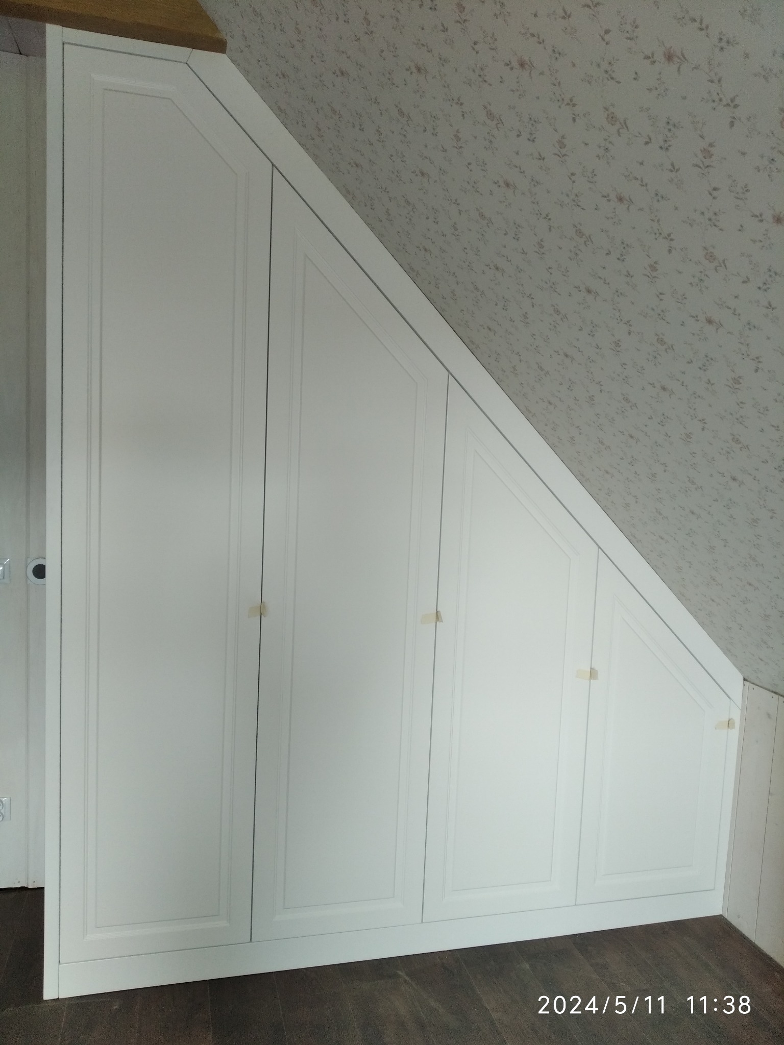 Biała zabudowa szafy wnękowej pod skosem na poddaszu, z widocznymi elementami maskującymi połączenia drzwiczek, na tle ściany z tapetą w drobne kwiaty.