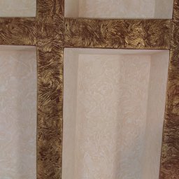 Jan & Co - Narożny fragment ściany z wnękami wykończonymi dekoracyjnym tynkiem w odcieniach beżu i złota, z fakturą przypominającą wirujące smugi.