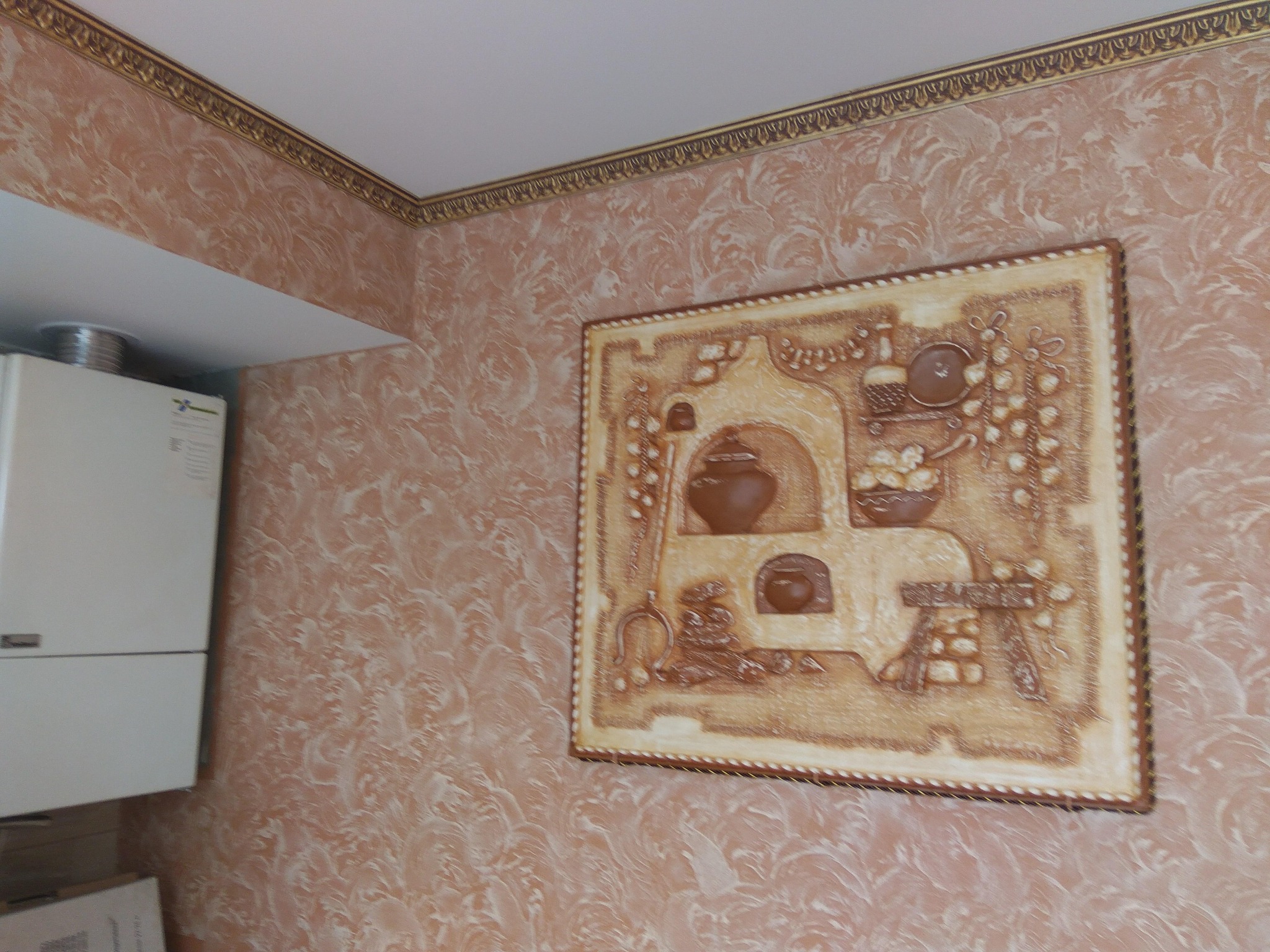 Wzorzysta ściana w odcieniach beżu i brązu z dekoracyjną listwą pod sufitem, na której wisi płaskorzeźba przedstawiająca rustykalną kuchnię z piecem, obok biały piec gazowy.