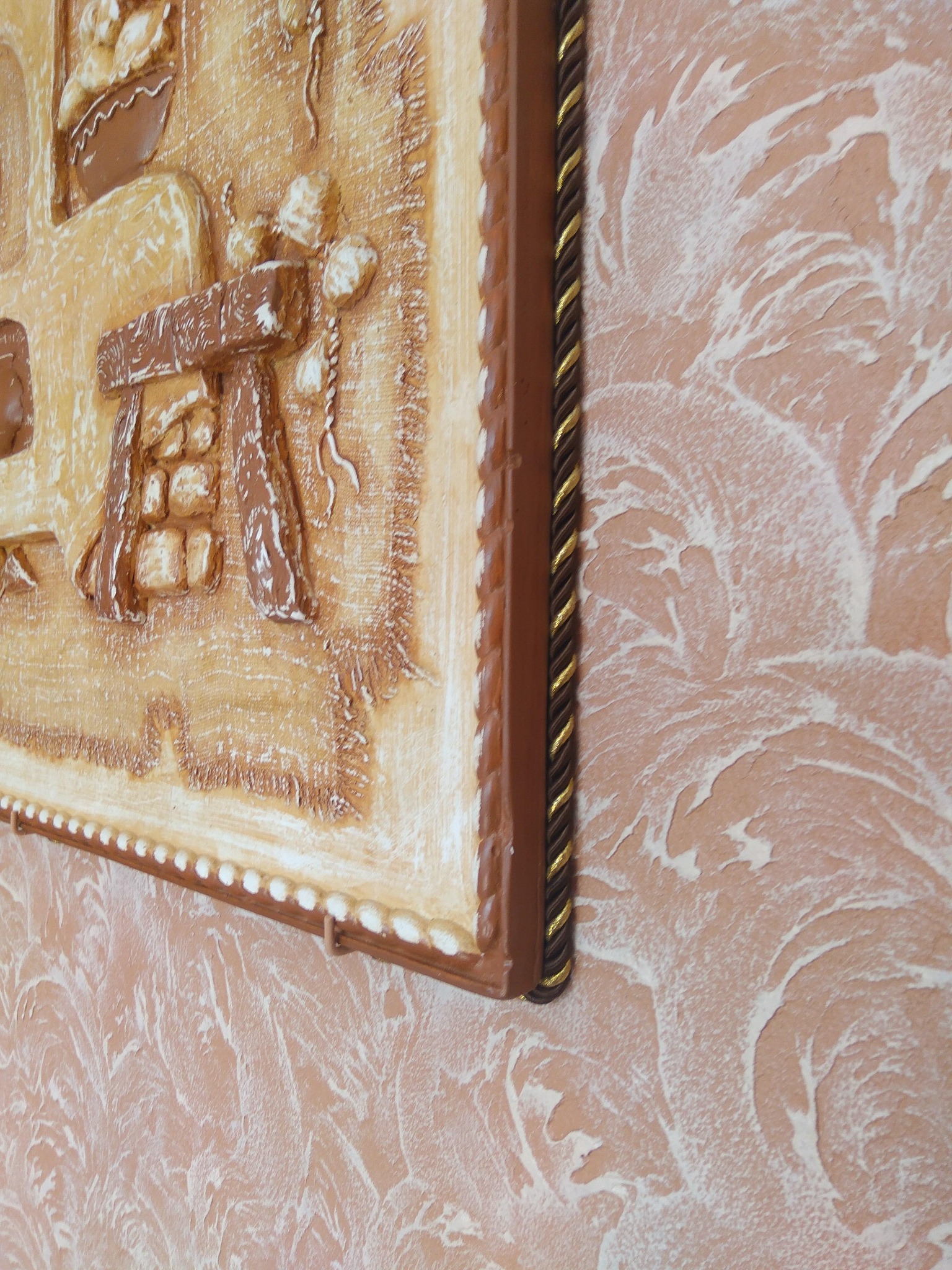 Dekoracyjna płaskorzeźba przedstawiająca scenę z wiejskiej kuchni, umieszczona na ścianie z efektem strukturalnym w odcieniach beżu i bieli, oprawiona w brązową ramę z akcentami złota.