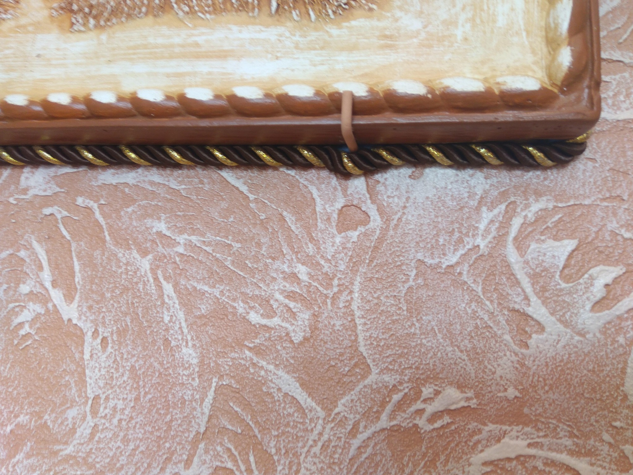 Fragment ściany z dekoracyjną strukturą w odcieniach brzoskwini i bieli, poniżej zdobiona rama z ornamentem sznurkowym w kolorze brązu i złota.