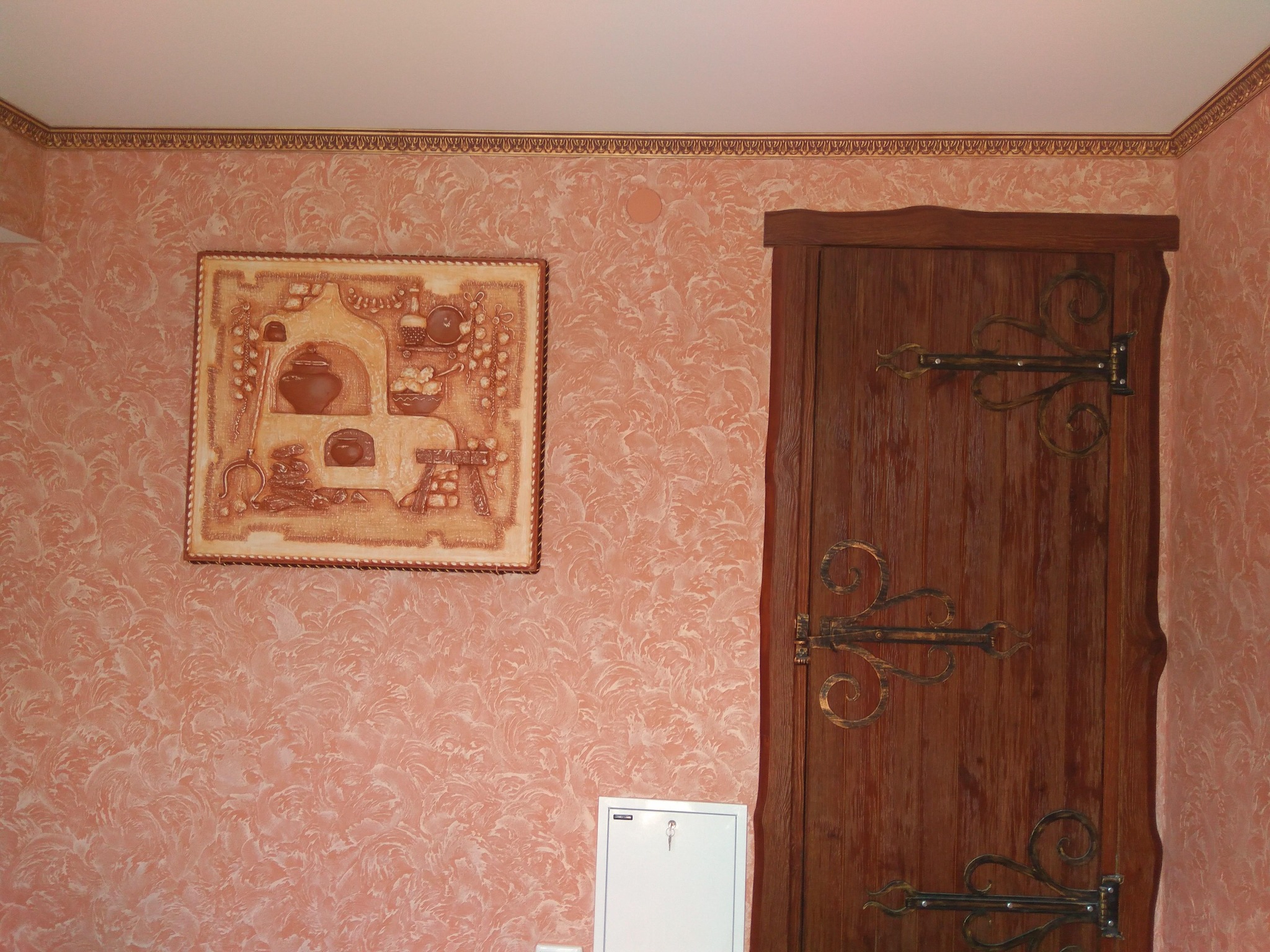 Ściana z dekoracyjnym tynkiem w odcieniach różu i beżu, ozdobiona obrazem z motywem pieca i drewnianymi drzwiami z metalowymi zawiasami w stylu retro.