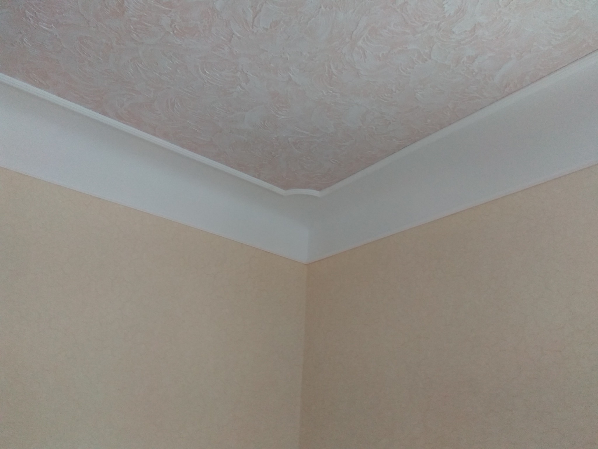 Róg pokoju z beżowymi ścianami o delikatnej strukturze, ozdobnym sufitem w odcieniach różu i białym wykończeniem sufitowym.