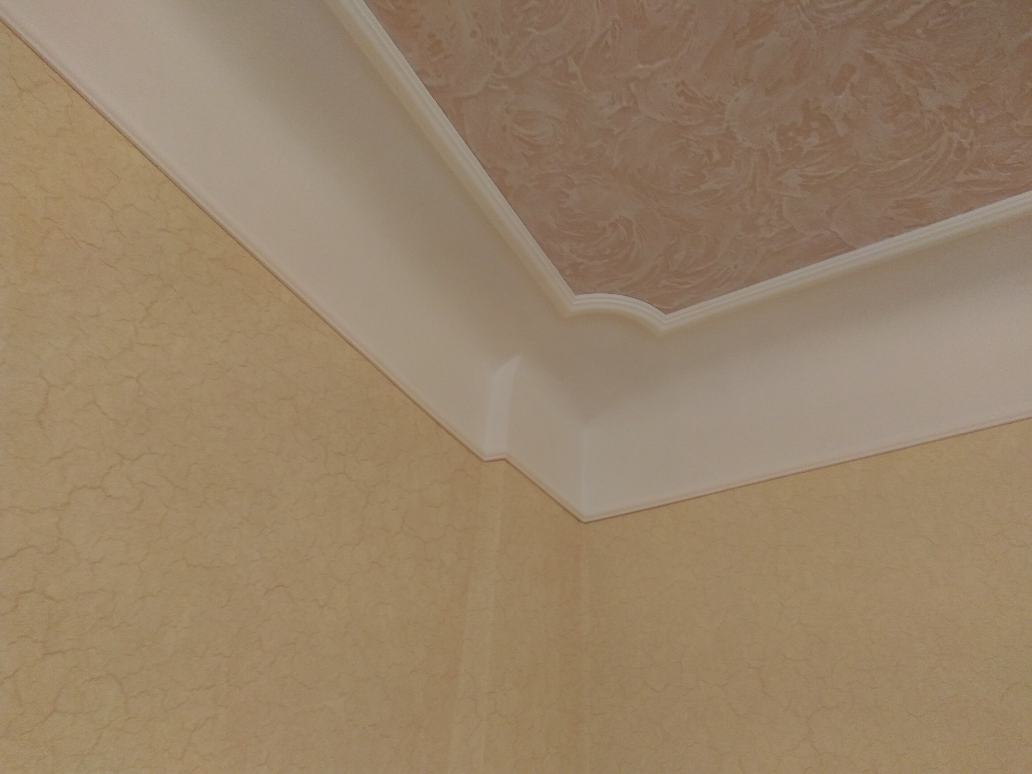 Wykończony narożnik pokoju z beżową ścianą o fakturze spękań i sufitem z dekoracyjnym wzorem, oddzielonym białym, profilowanym gzymsem.