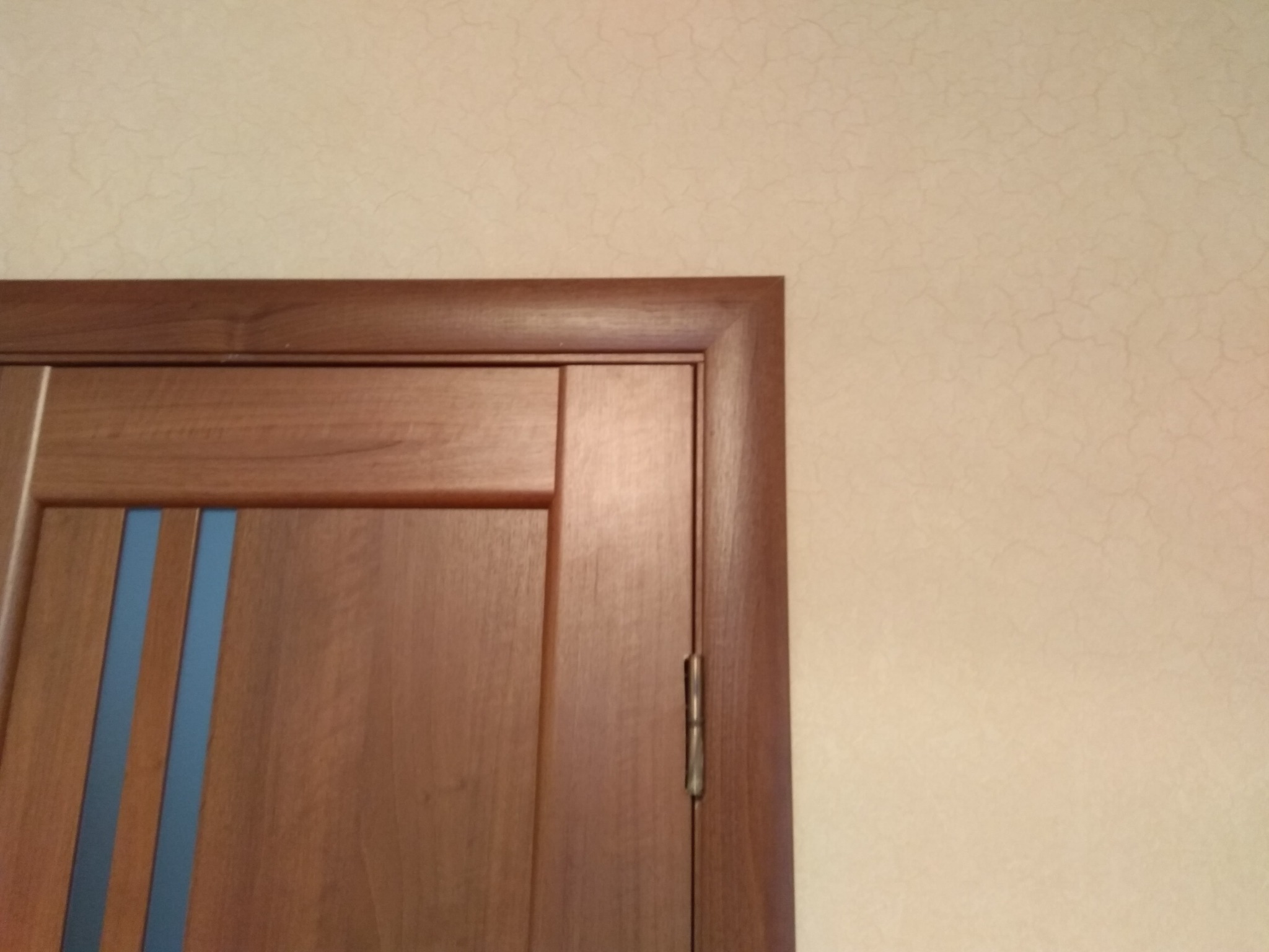 Fragment drewnianych drzwi wewnętrznych z trzema pionowymi, niebieskimi wstawkami, zawias w kolorze starego złota, kremowa ściana w tle z delikatną fakturą.