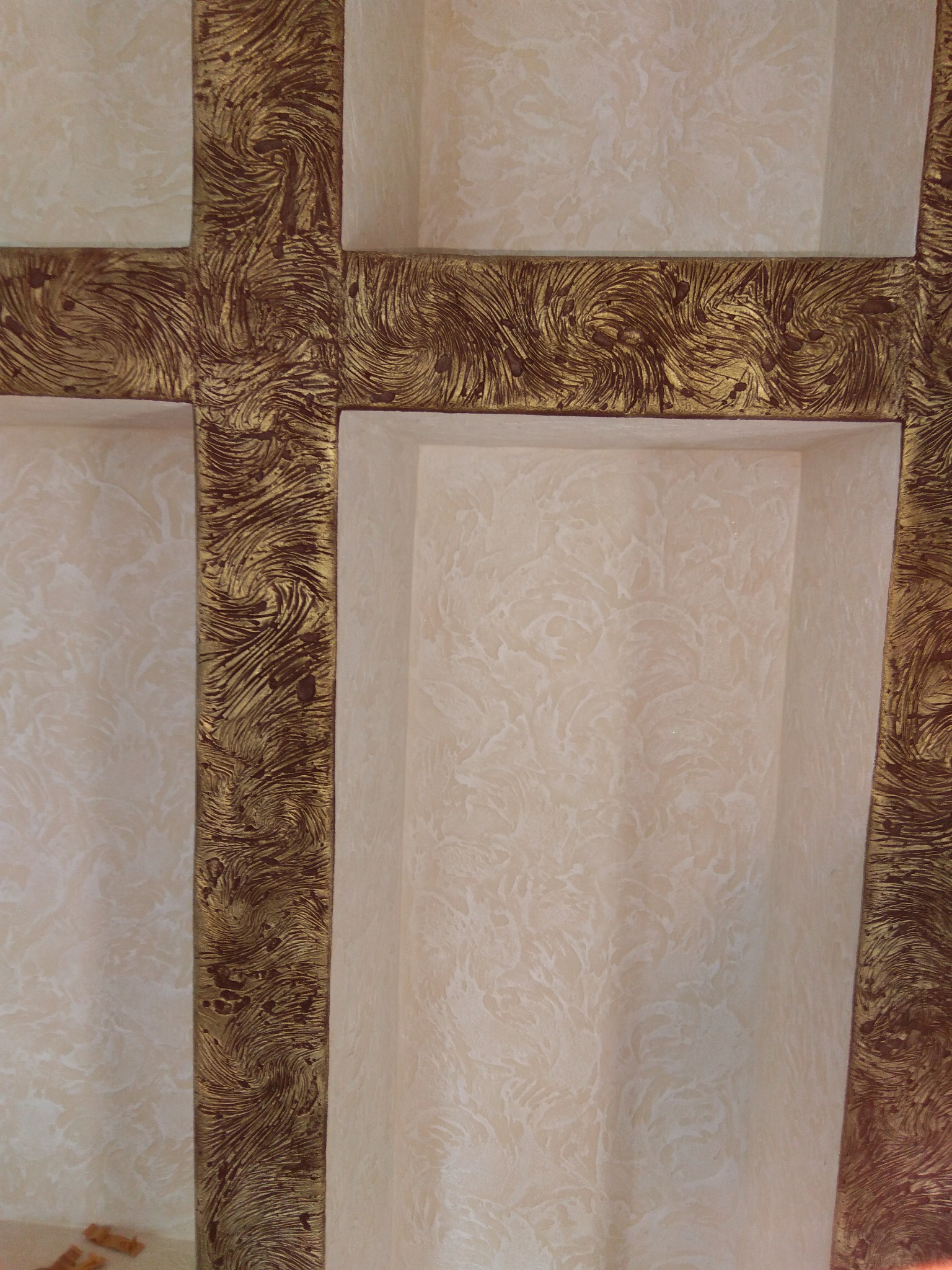 Narożny fragment ściany z wnękami wykończonymi dekoracyjnym tynkiem w odcieniach beżu i złota, z fakturą przypominającą wirujące smugi.