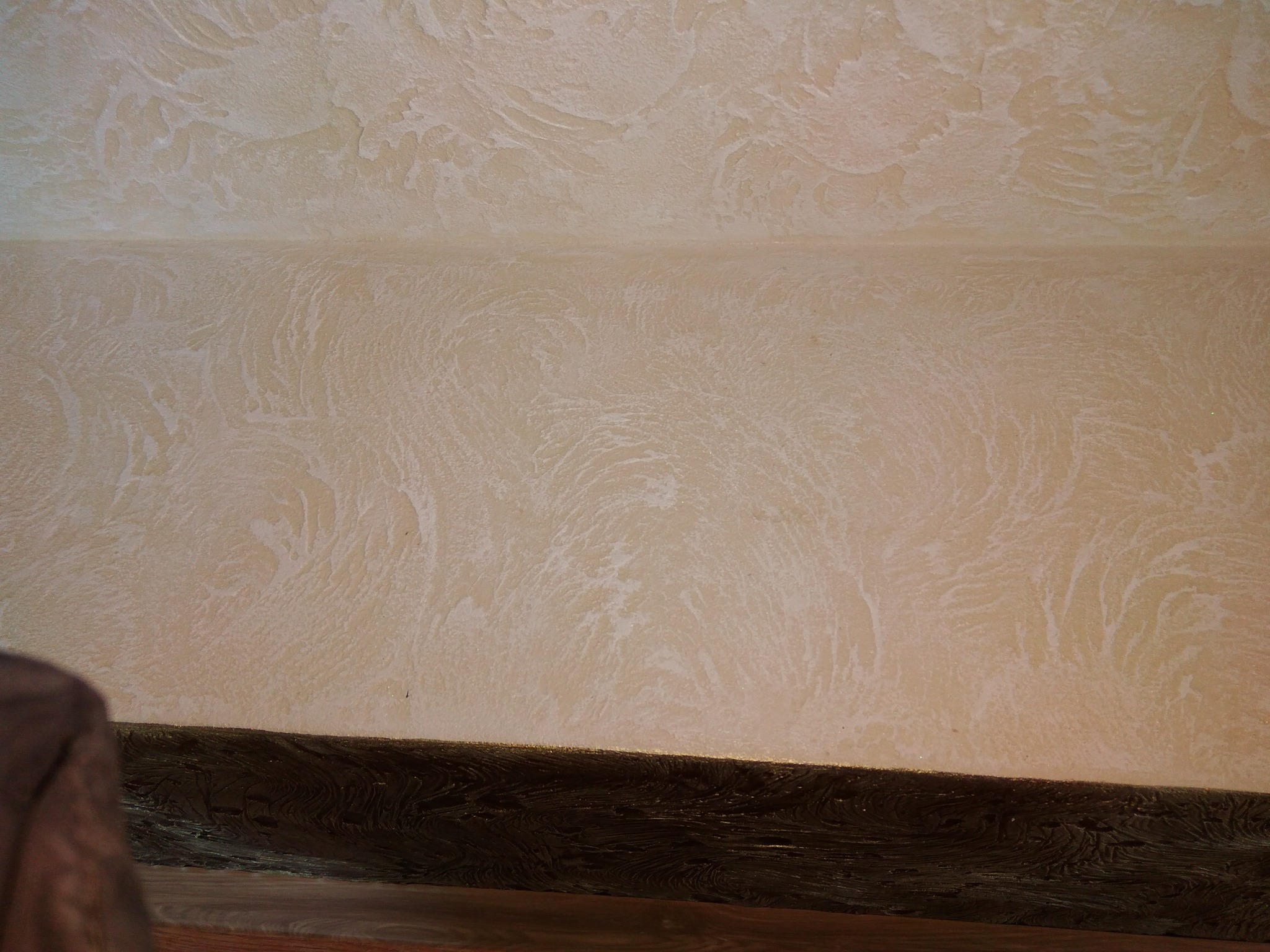 Beżowa ściana z dekoracyjną fakturą imitującą fale, z ciemnobrązową listwą przypodłogową i fragmentem drewnianej podłogi.