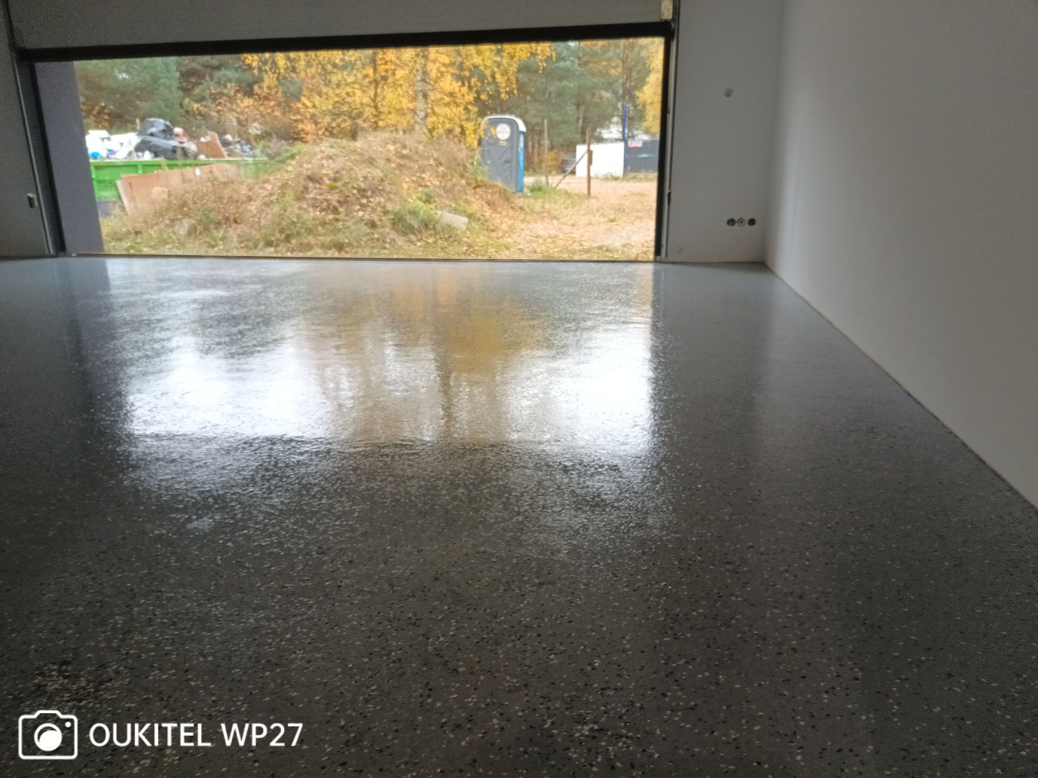 Świeżo wylana, błyszcząca, szara posadzka żywiczna z widocznymi drobnymi ziarnami, odbijająca światło w pustym pomieszczeniu z otwartą bramą garażową i widokiem na jesienne drzewa, kontener...