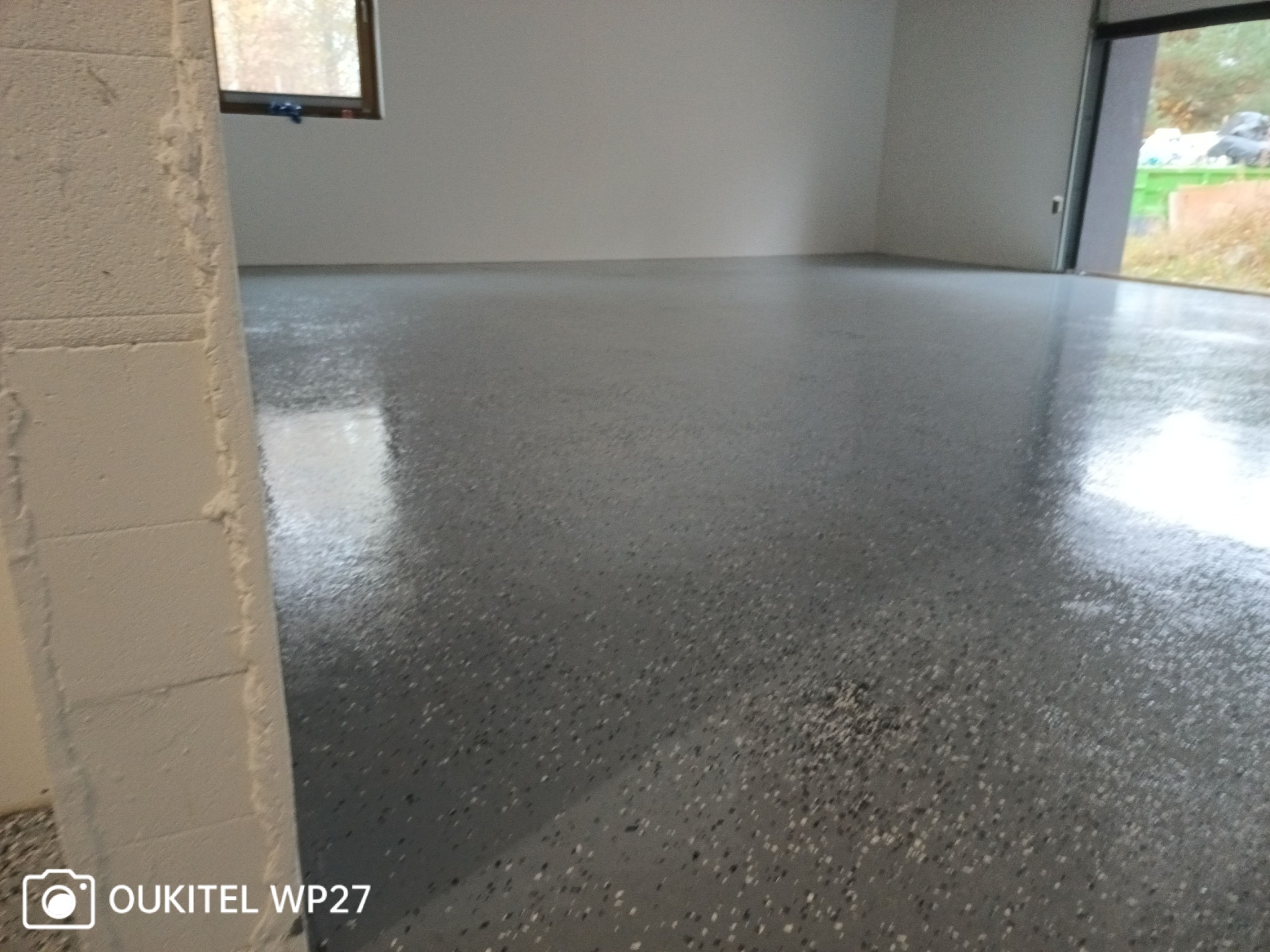 Błyszcząca, szara posadzka żywiczna z ciemnymi inkluzjami w pustym pomieszczeniu z oknem i otwartymi drzwiami garażowymi, widok z niskiego kąta.