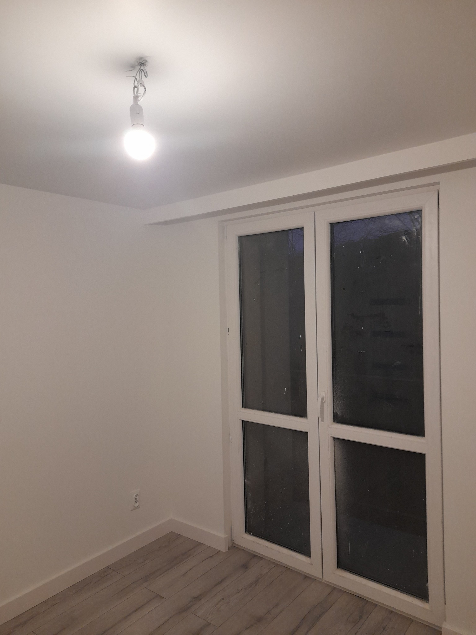 Pusty pokój po remoncie z panelami podłogowymi, białymi ścianami i dwuskrzydłowymi drzwiami balkonowymi; widoczna surowa żarówka na kablu.