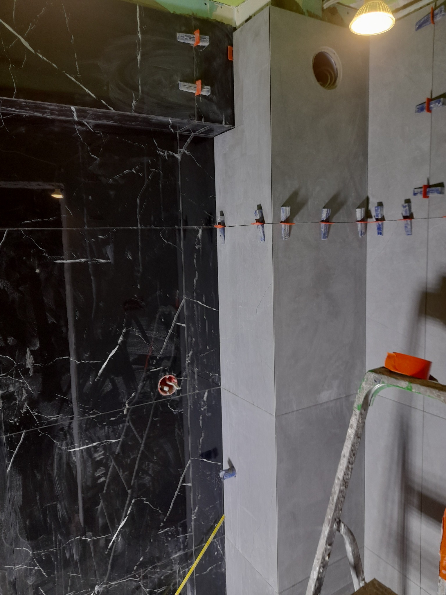 Świeżo ułożone, czarne płytki imitujące marmur na ścianie, kontrastujące z szarymi płytkami. Widoczne klipsy poziomujące i sznurki, drabina z pomarańczowym wiadrem na górze, surowa żarówka...