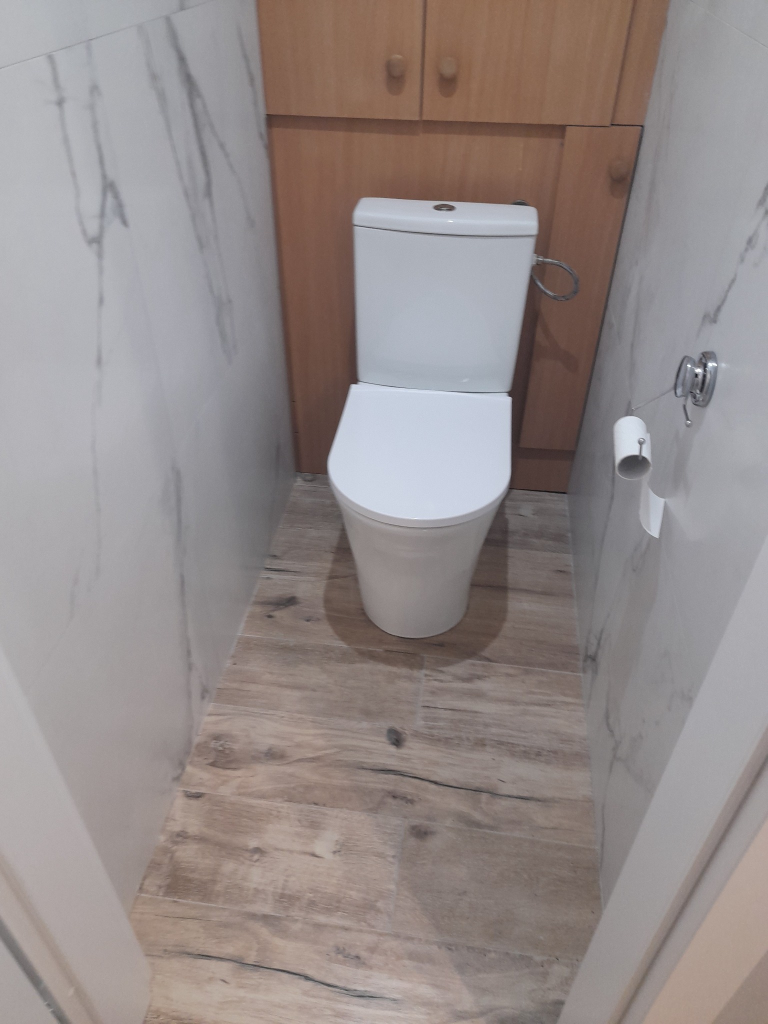 Biała toaleta kompaktowa w małej łazience z imitacją drewnianej podłogi, marmurowe płytki na ścianach i szafka wisząca z jasnego drewna.