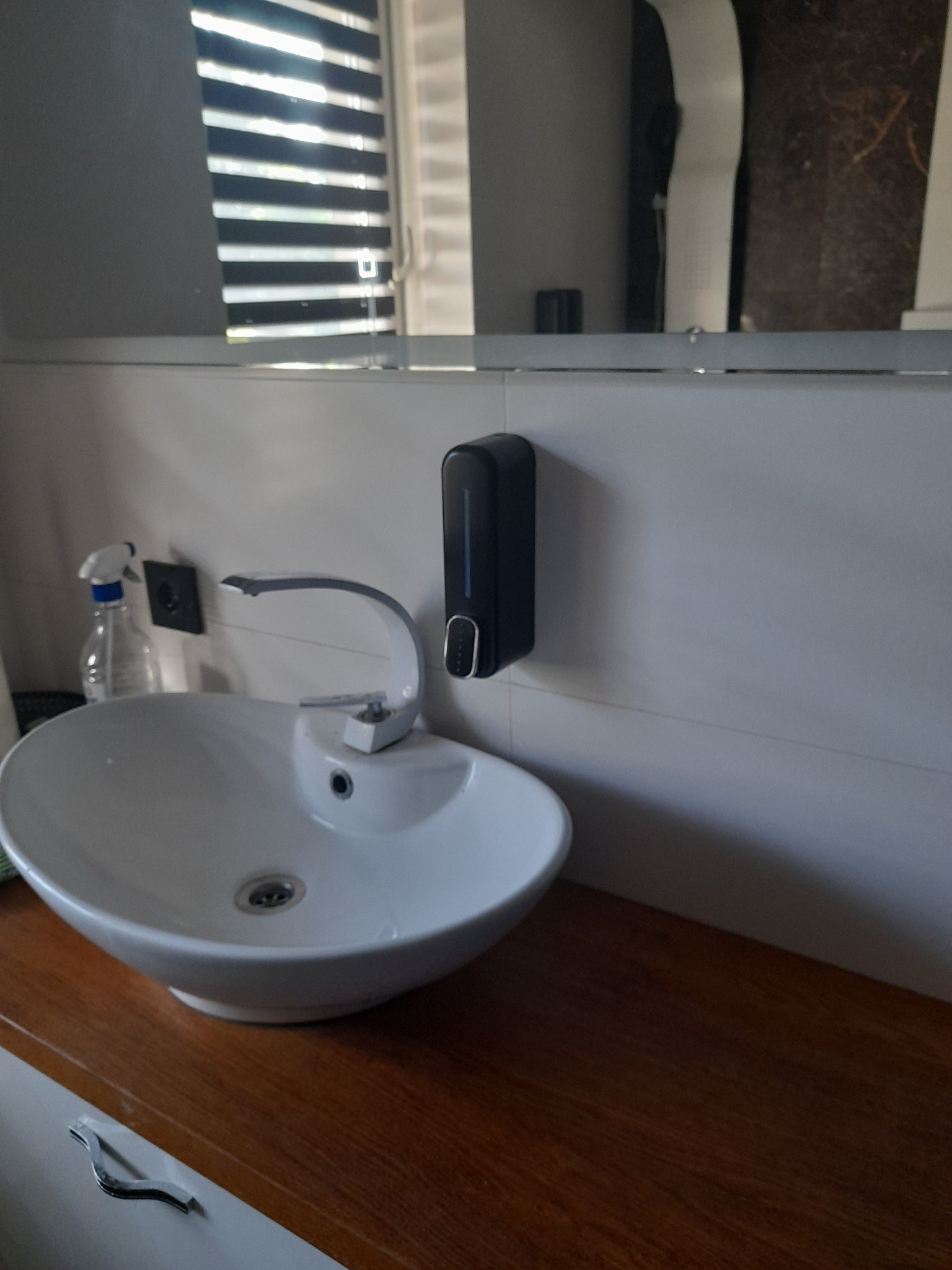 Nowoczesna umywalka nablatowa z chromowaną baterią, zamontowana na blacie z ciemnego drewna, z dozownikiem mydła w kolorze czarnym na ścianie wyłożonej białymi płytkami. W tle widoczne lustro...