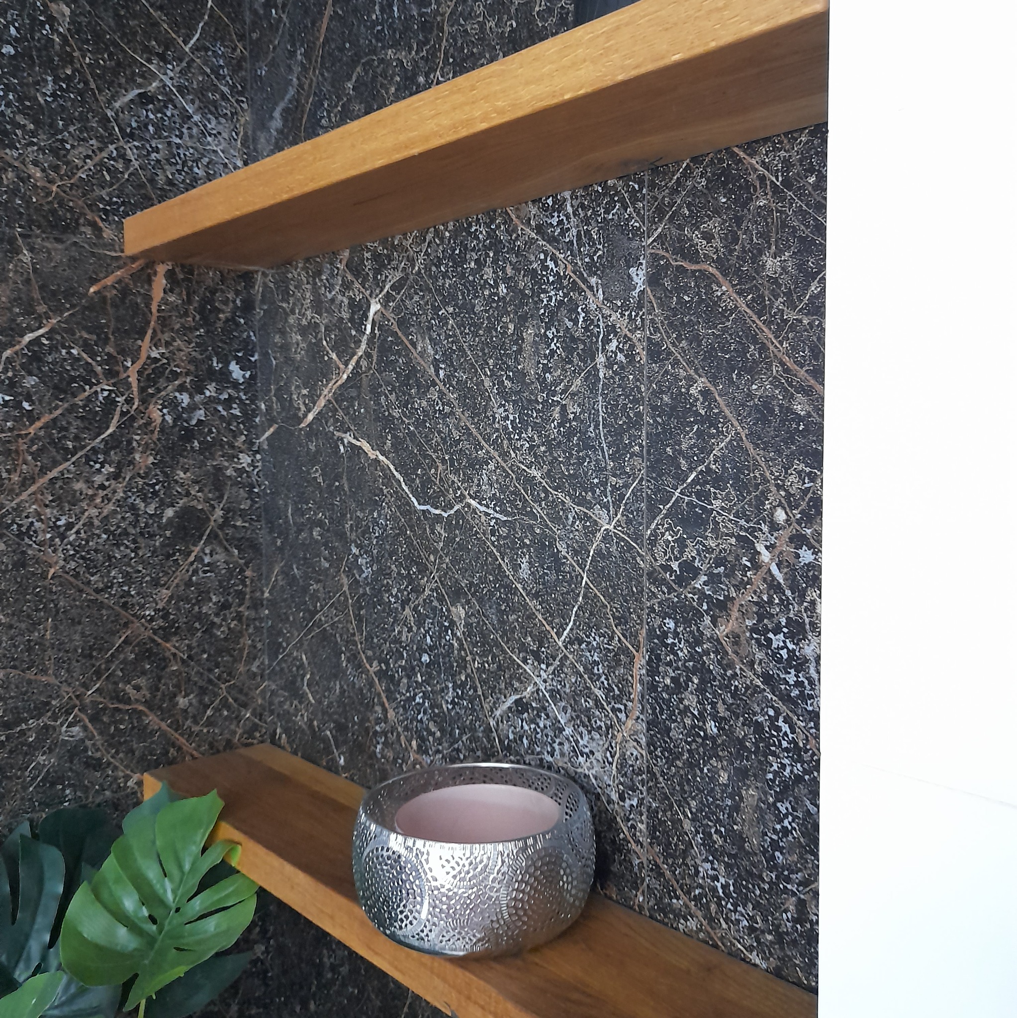 Dwie drewniane półki na tle ściany z czarnego marmuru z białymi i złotymi żyłami. Na dolnej półce stoi srebrny świecznik z ażurowym wzorem, obok widoczne liście sztucznej rośliny.