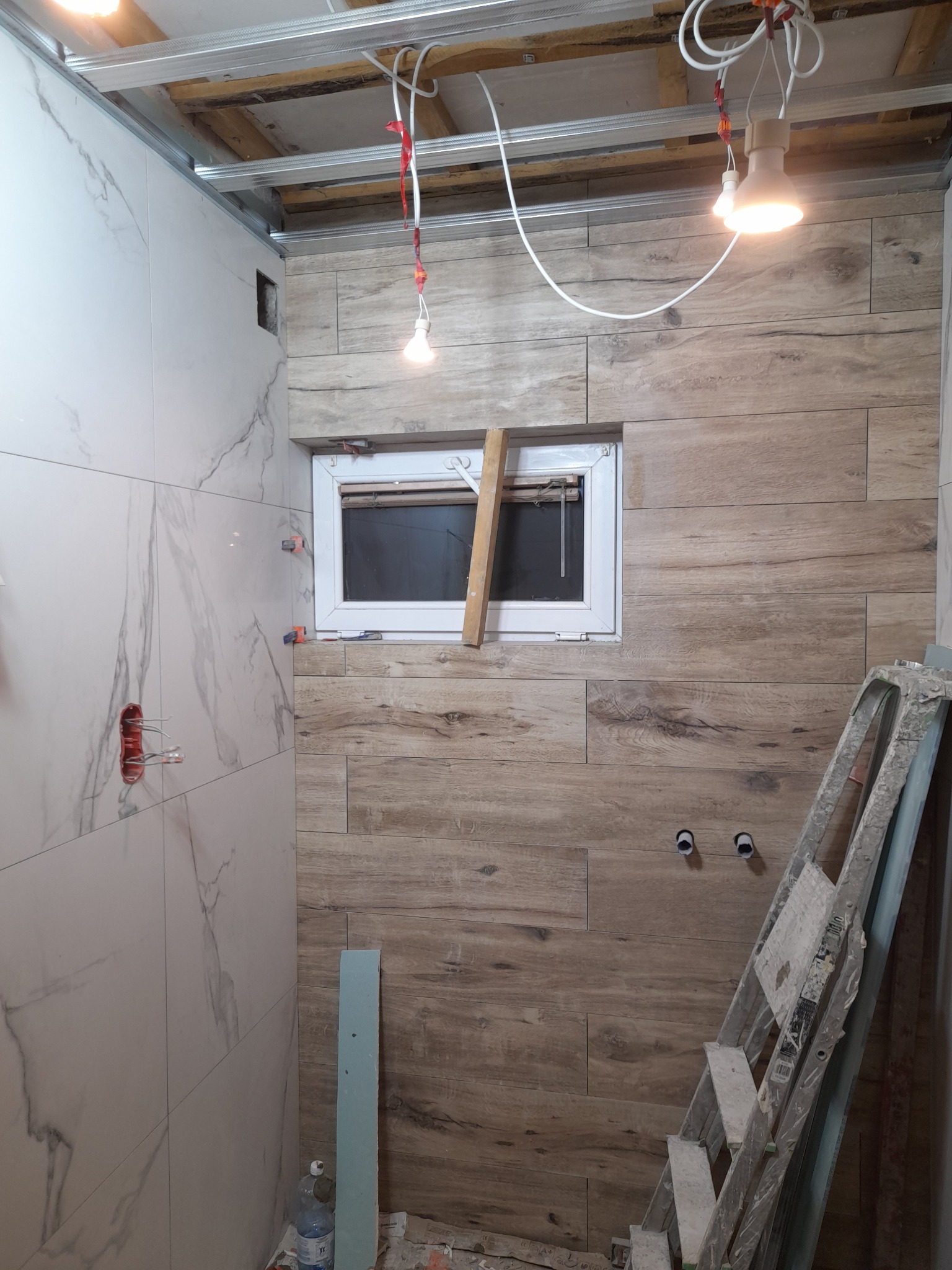 Łazienka w trakcie remontu: białe marmurowe płytki na ścianie obok instalacji elektrycznej, płytki imitujące drewno na ścianie z oknem zabezpieczonym deską, drabina oparta o ścianę, surowy sufit...