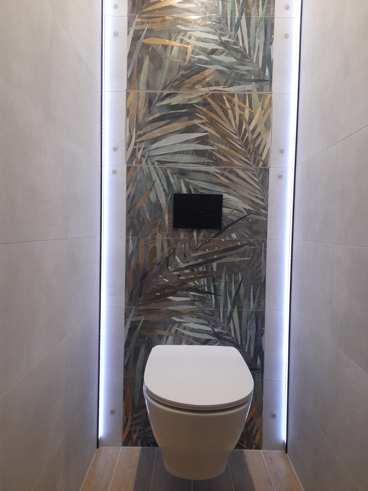 Nowoczesna toaleta podwieszana z podświetlanymi płytkami dekoracyjnymi z motywem liści palmowych i szarymi płytkami ściennymi.