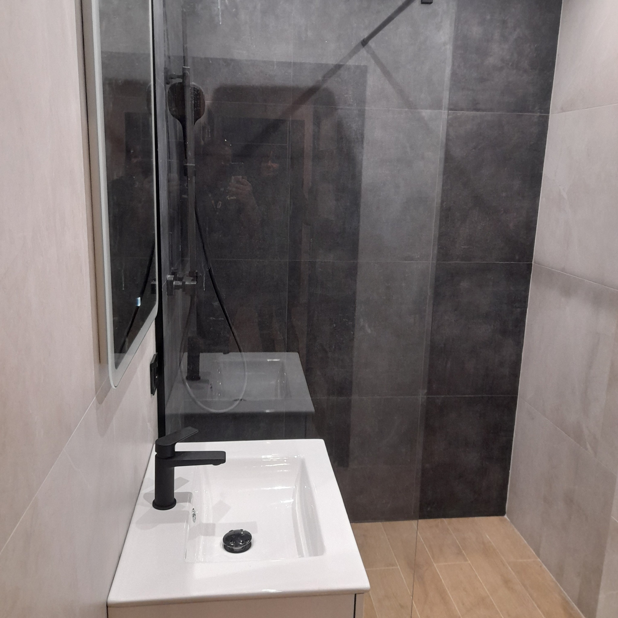 Nowoczesna łazienka z kabiną prysznicową typu walk-in, umywalką nablatową z czarną armaturą i dużym lustrem, wykończona w odcieniach szarości i beżu z akcentem drewnopodobnych płytek na podłodze.