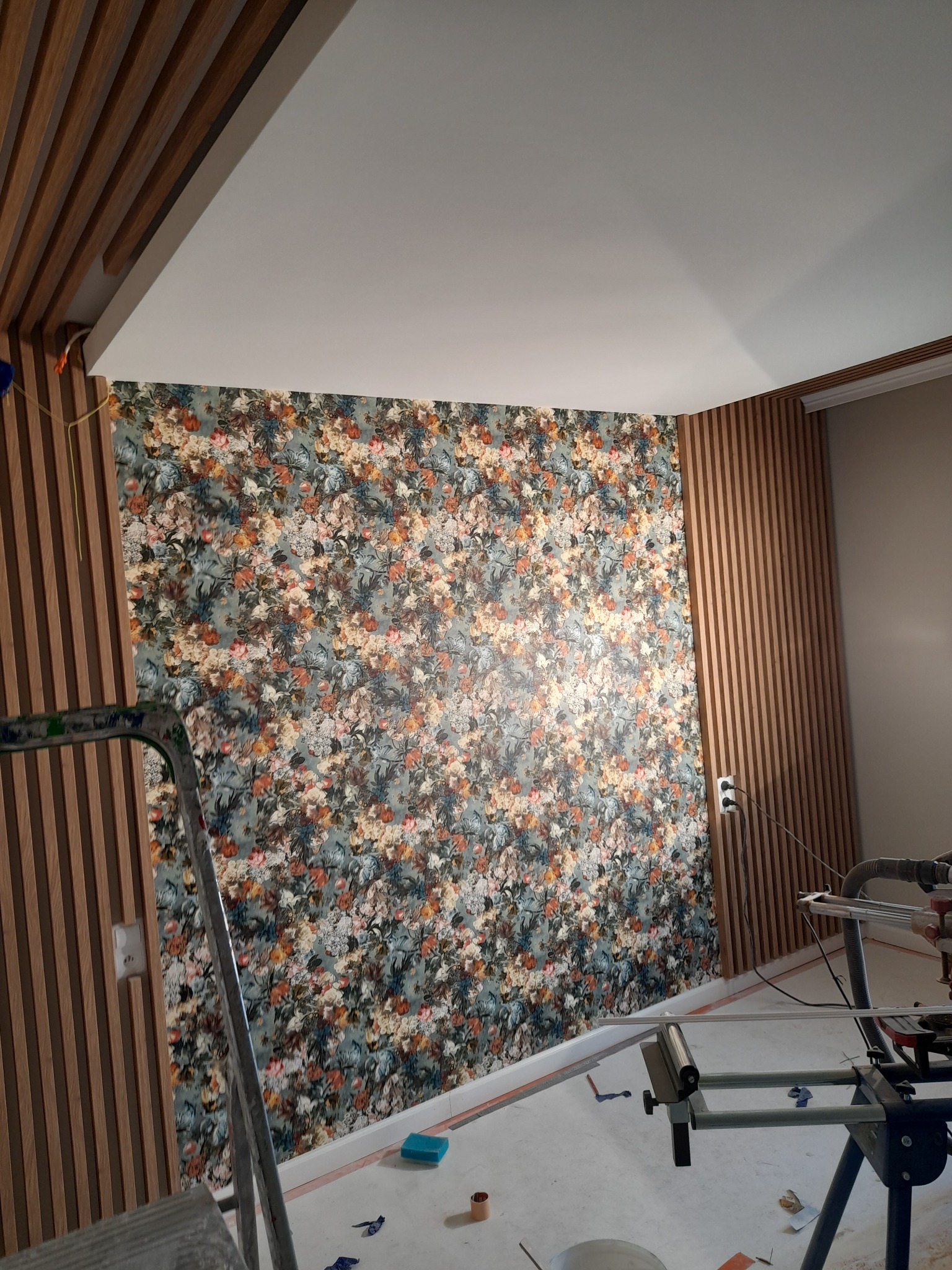 Ściana z tapetą w kwiaty i drewnianymi lamelami; widoczny fragment drabiny i narzędzi, w trakcie prac wykończeniowych.
