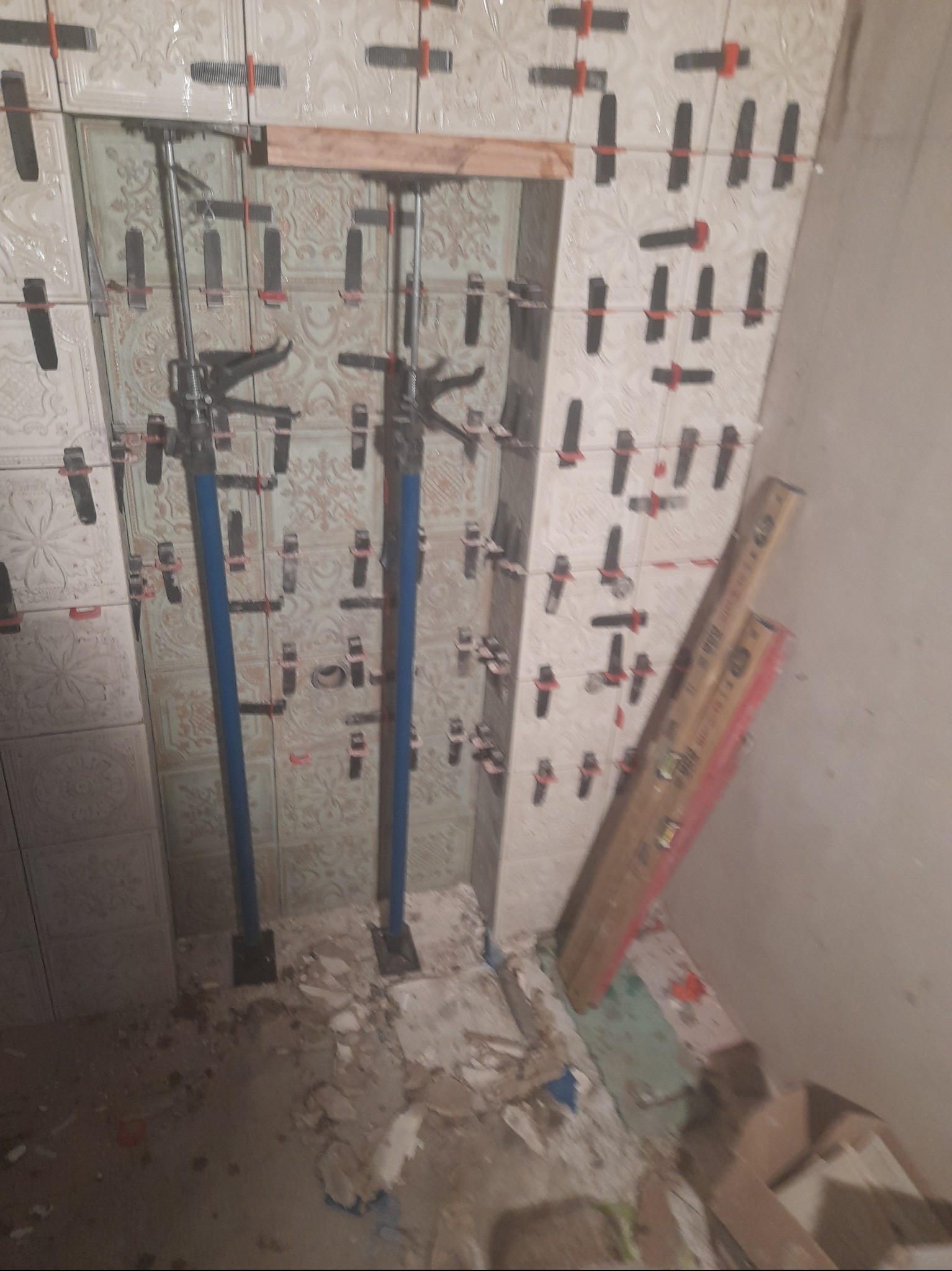 Ściana łazienki w trakcie układania płytek z użyciem systemu poziomowania, widoczne podpory teleskopowe i poziomica, na podłodze resztki zaprawy.