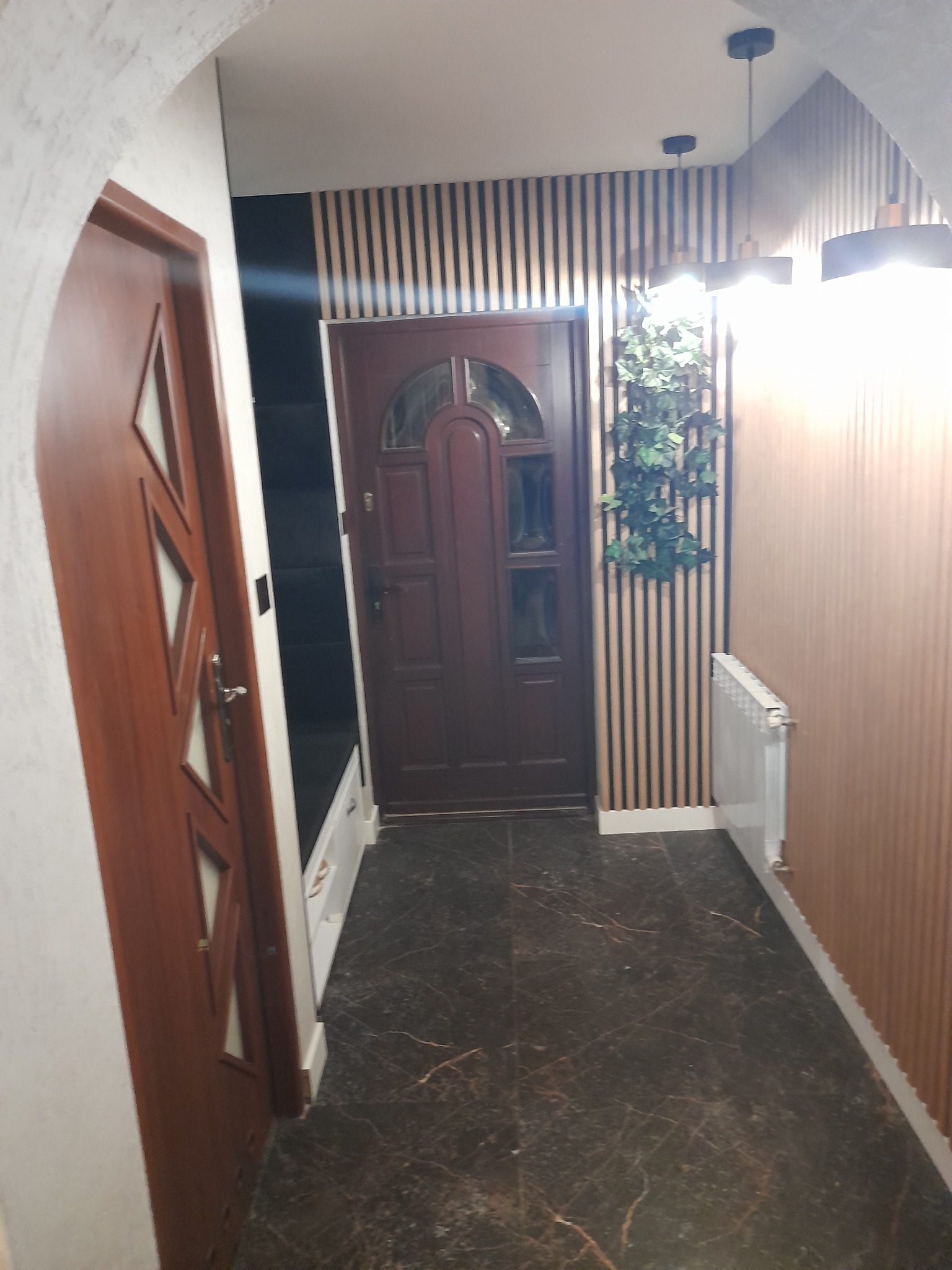 Korytarz z ciemną podłogą imitującą marmur, drewnianą boazerią lamelową na ścianach, drzwiami wewnętrznymi z dekoracyjnymi przeszkleniami i zewnętrznymi w kolorze ciemnego drewna, zabudowaną szafką...