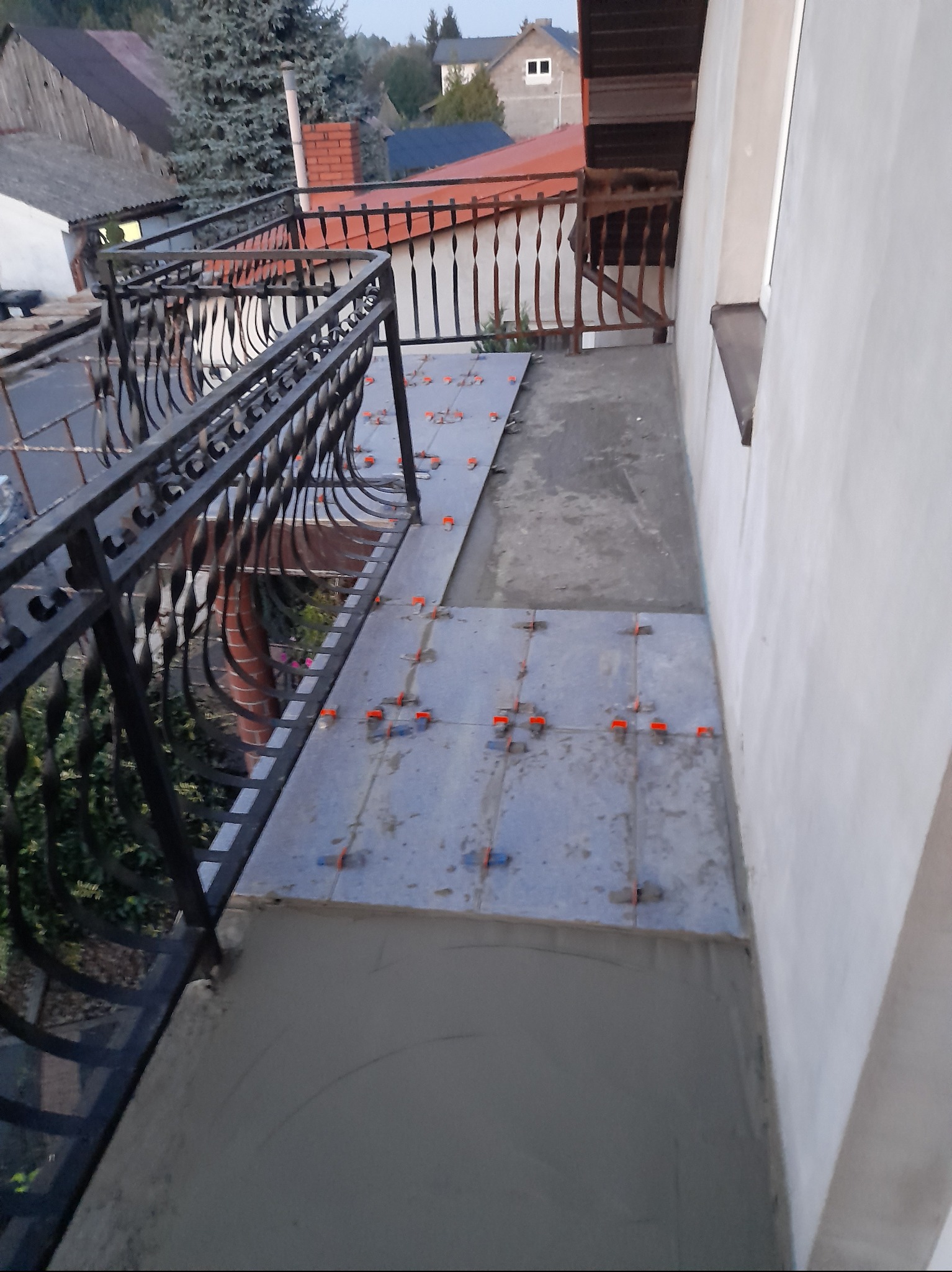 Układanie szarych płytek na balkonie z czarną, kutą balustradą, widoczne pomarańczowe krzyżyki dystansowe, częściowo położone płytki na zaprawie, w tle zabudowa jednorodzinna.