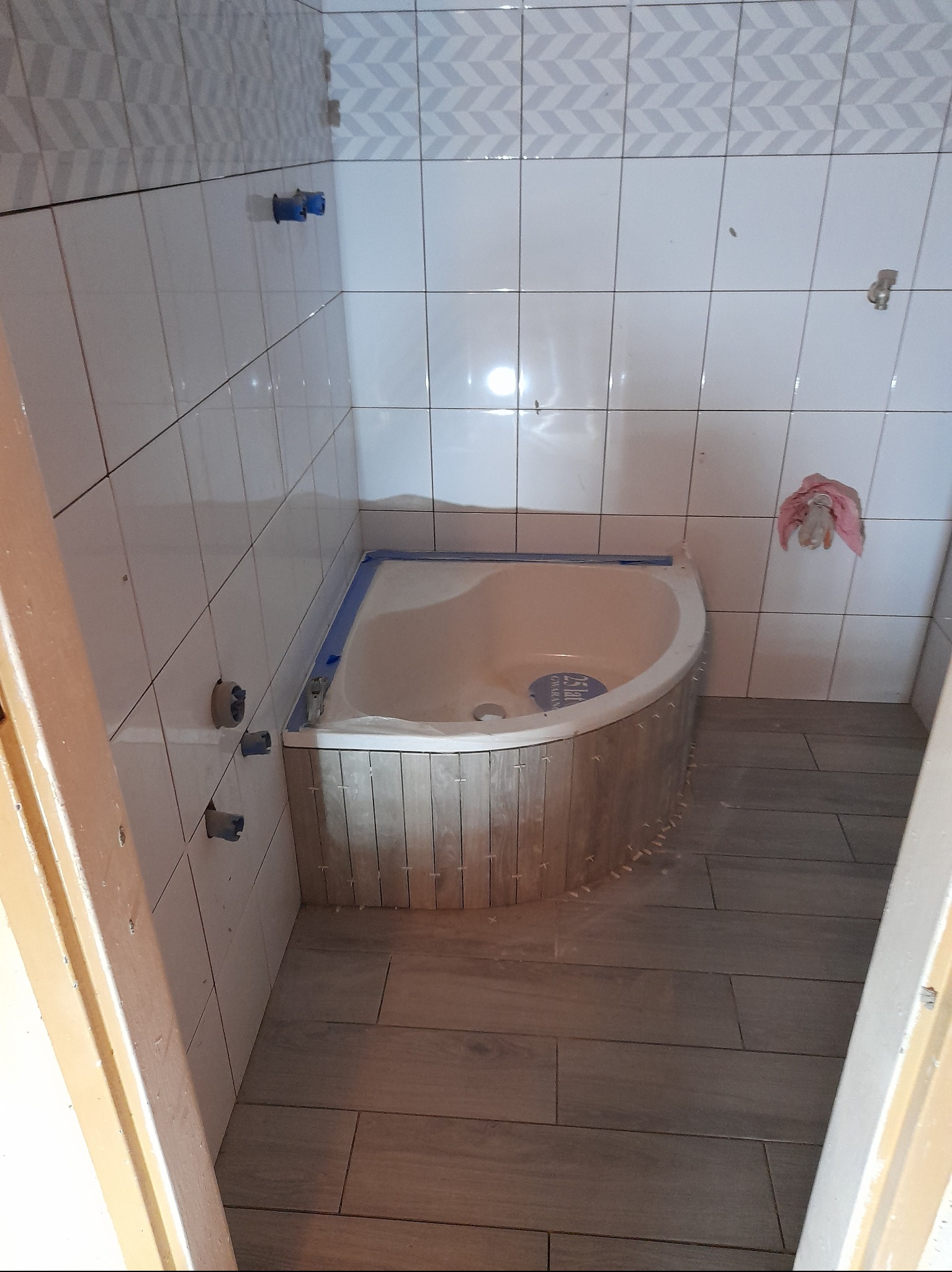 Narożna wanna z obudową imitującą drewno, zabezpieczona niebieską taśmą malarską, w łazience z ułożonymi płytkami podłogowymi przypominającymi panele; widoczne krzyżyki dystansowe przy wannie i...