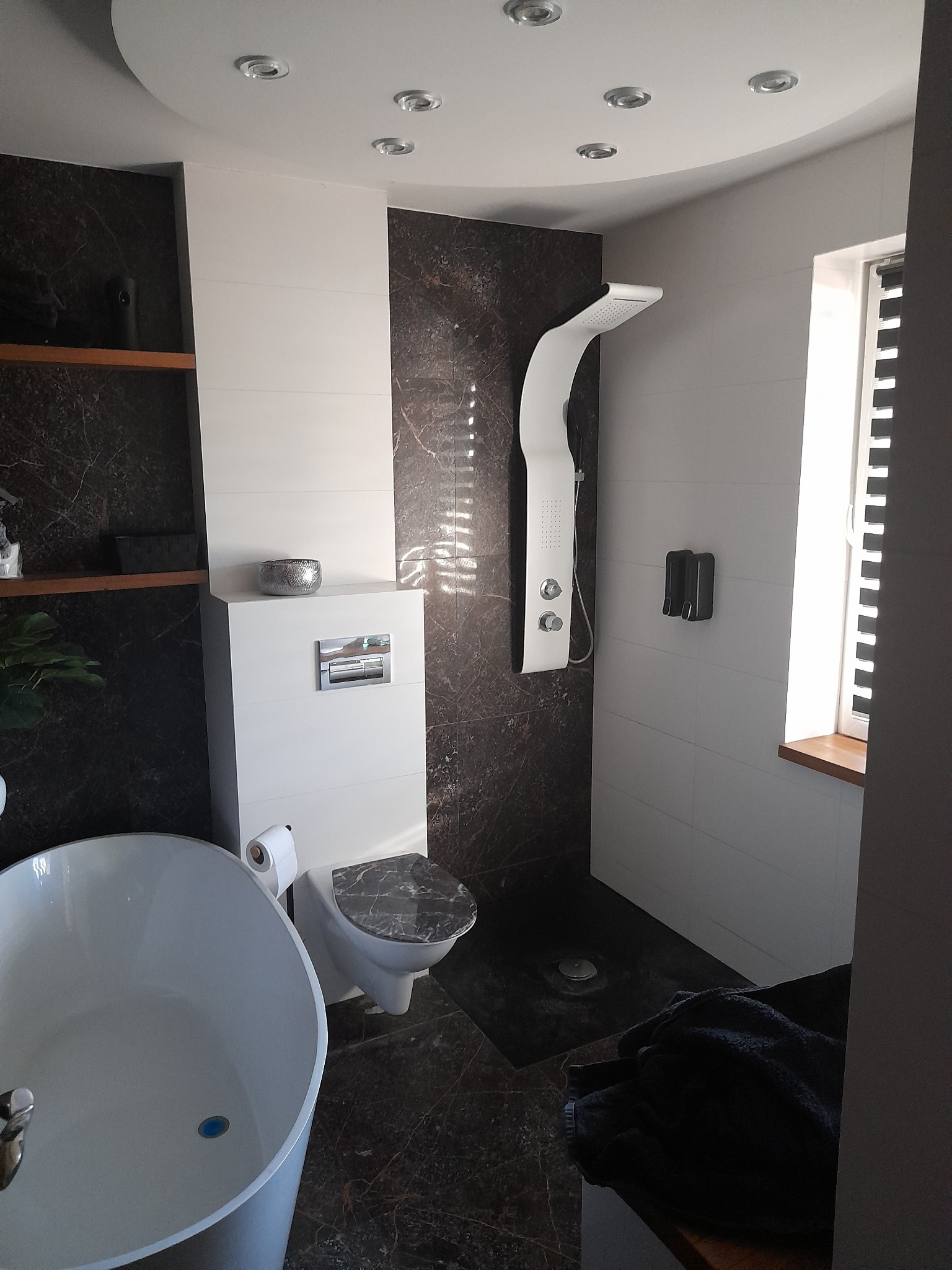 Nowoczesna łazienka z kabiną prysznicową typu walk-in, białą wanną wolnostojącą, podwieszaną toaletą z marmurową deską i ciemnymi płytkami imitującymi marmur na ścianach, oświetlenie punktowe...