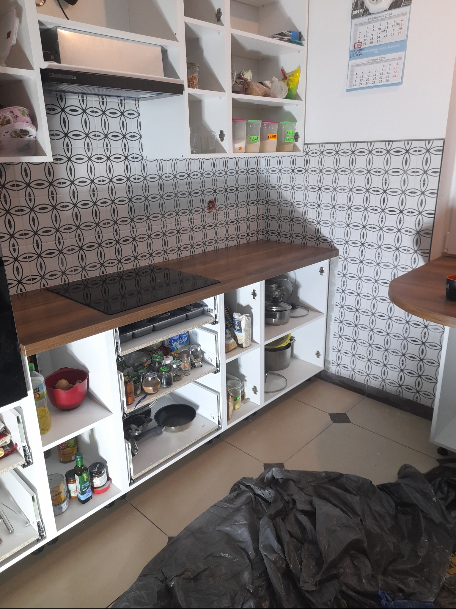 Fragment kuchni w trakcie prac wykończeniowych: widoczne białe szafki z drewnianym blatem, czarna płyta indukcyjna, ozdobne płytki na ścianie, kalendarz na ścianie i folia ochronna na podłodze.