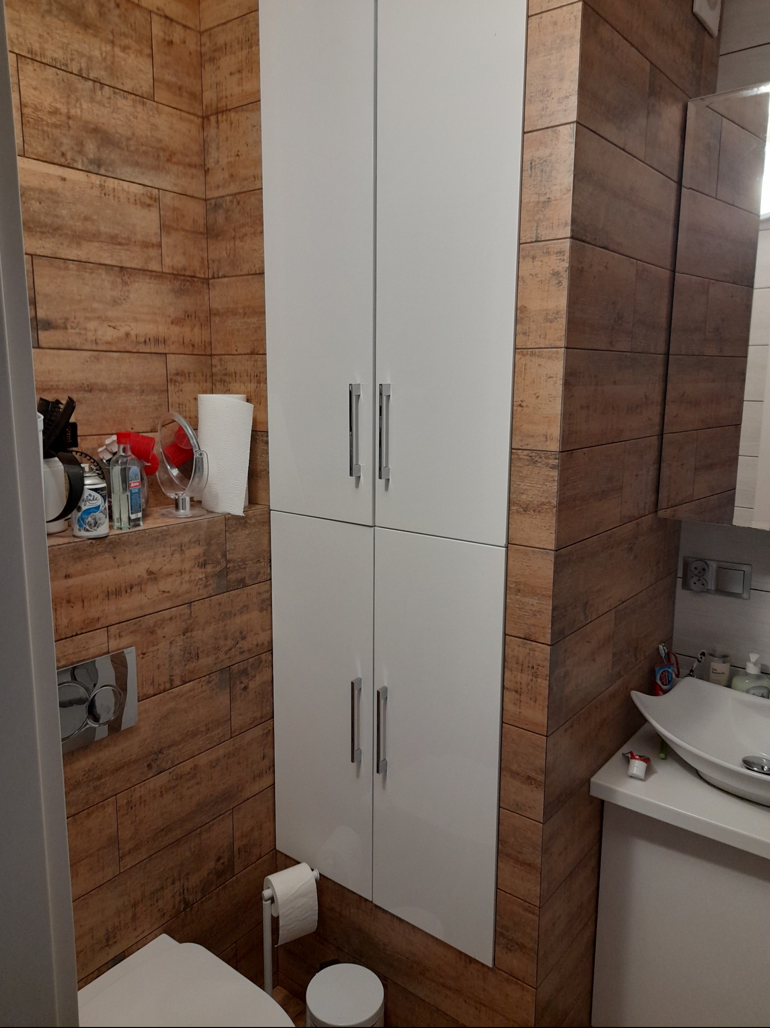 Biała szafka łazienkowa na tle ściany wyłożonej brązowymi płytkami imitującymi drewno, widoczny fragment toalety i umywalki.