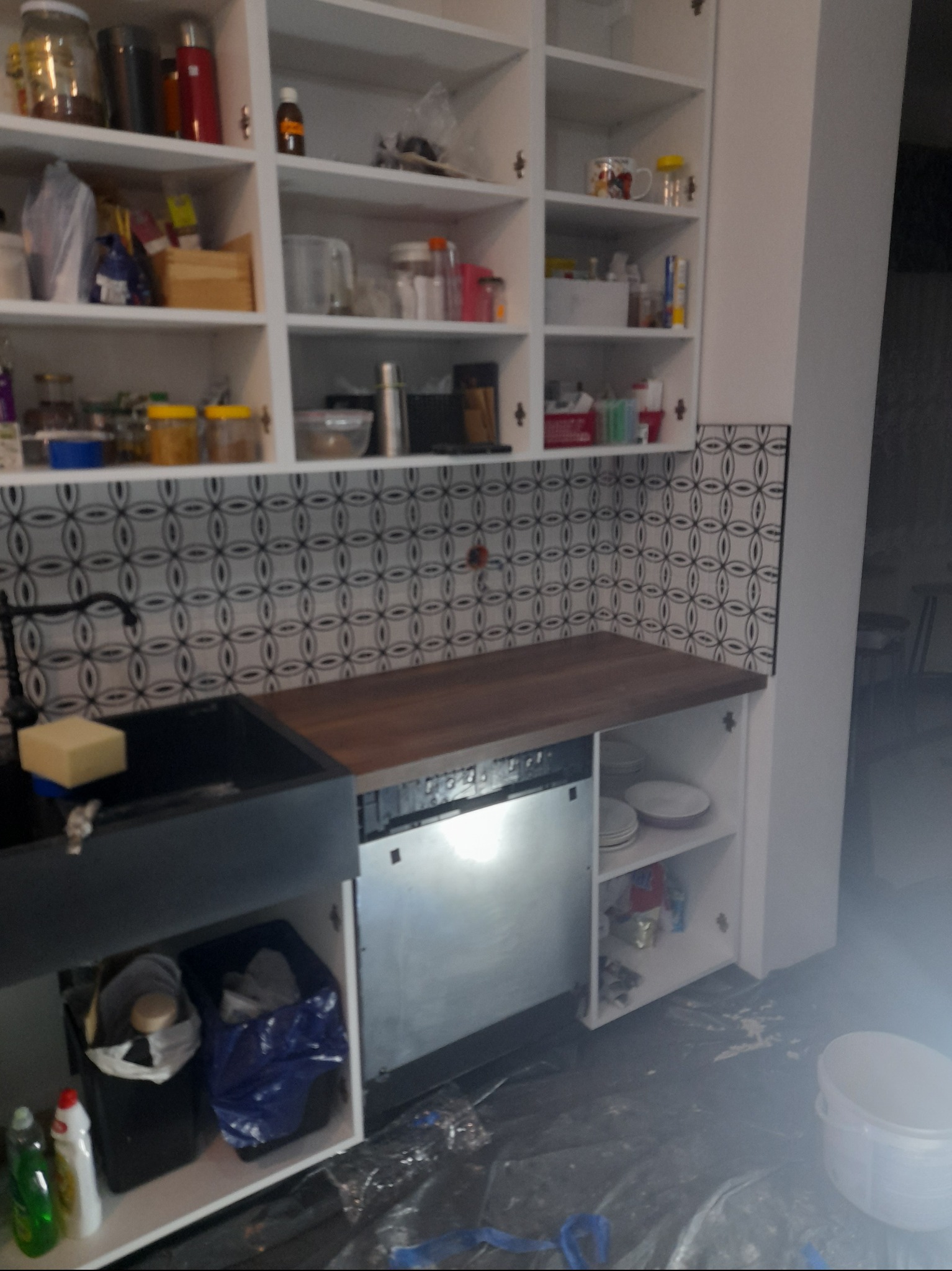 Fragment kuchni w trakcie prac: czarny zlew farmerski z baterią i gąbką, blat z ciemnego drewna, zabudowa kuchenna z białymi szafkami, wzorzyste płytki na ścianie, widoczna zmywarka i akcesoria...