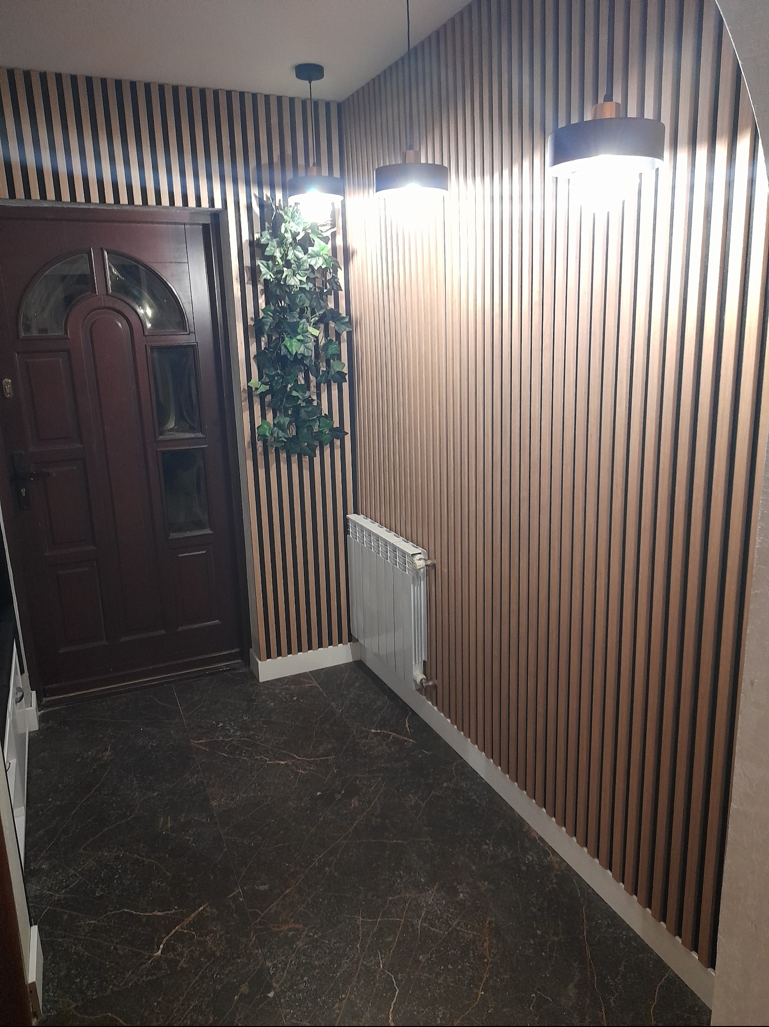 Przedpokój z drewnianymi lamelami na ścianach, ciemną, marmurkową podłogą, brązowymi drzwiami wejściowymi i rośliną pnącą się po ścianie, oświetlony trzema lampami wiszącymi.