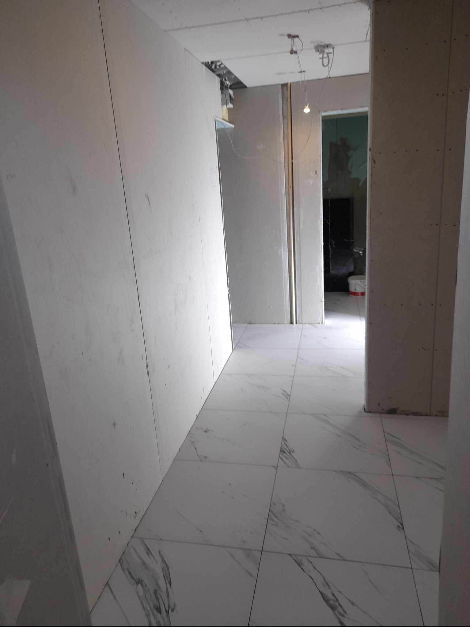 Korytarz w trakcie wykańczania: ściany z płyt karton gips, białe płytki podłogowe imitujące marmur z szarymi żyłami, widoczne instalacje elektryczne pod sufitem.