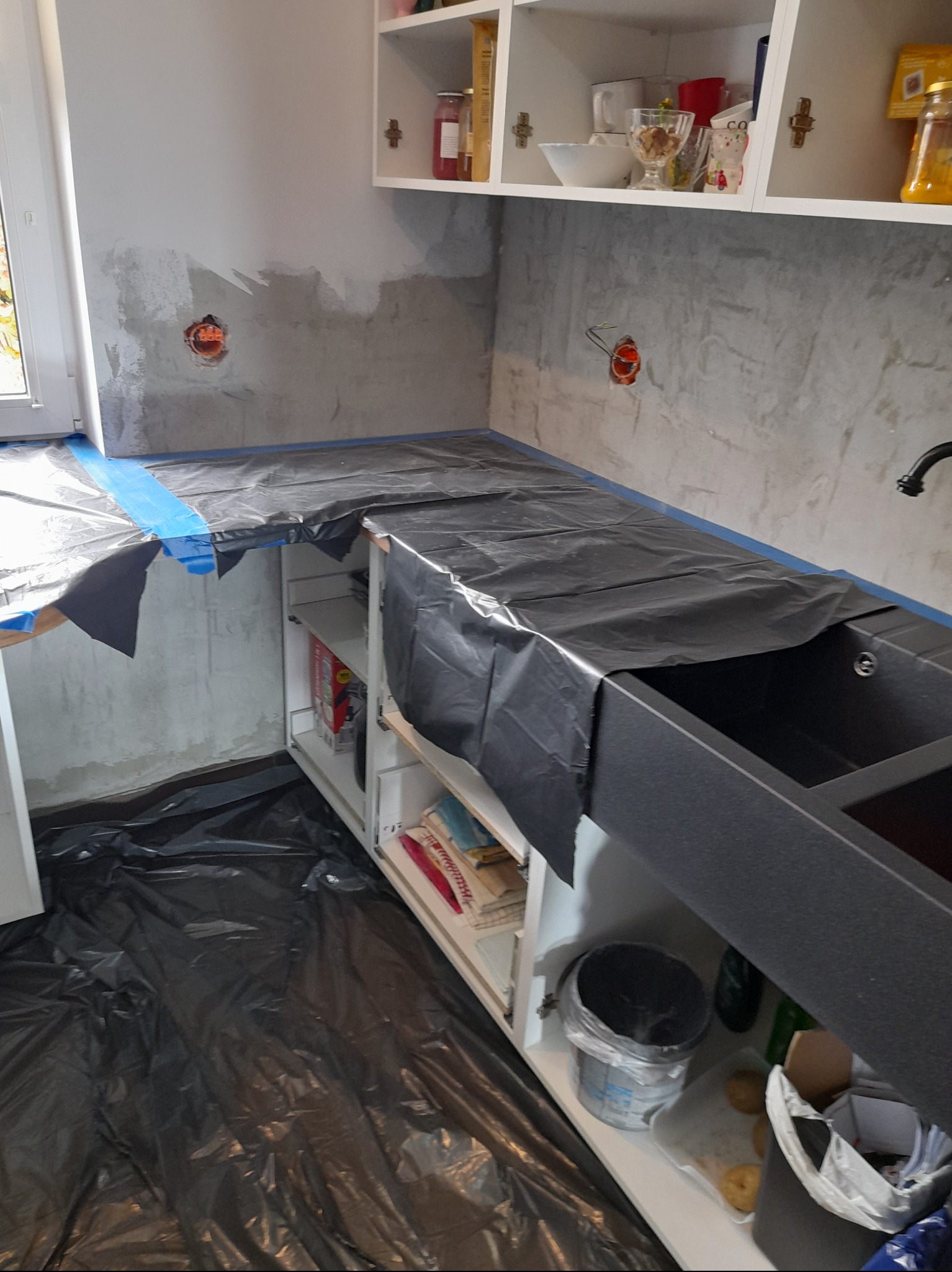 Przygotowanie do prac wykończeniowych w kuchni: zabezpieczona folią blat i podłoga, ściana częściowo przygotowana pod gładź z widocznymi puszkami elektrycznymi, białe szafki wiszące i stojące.