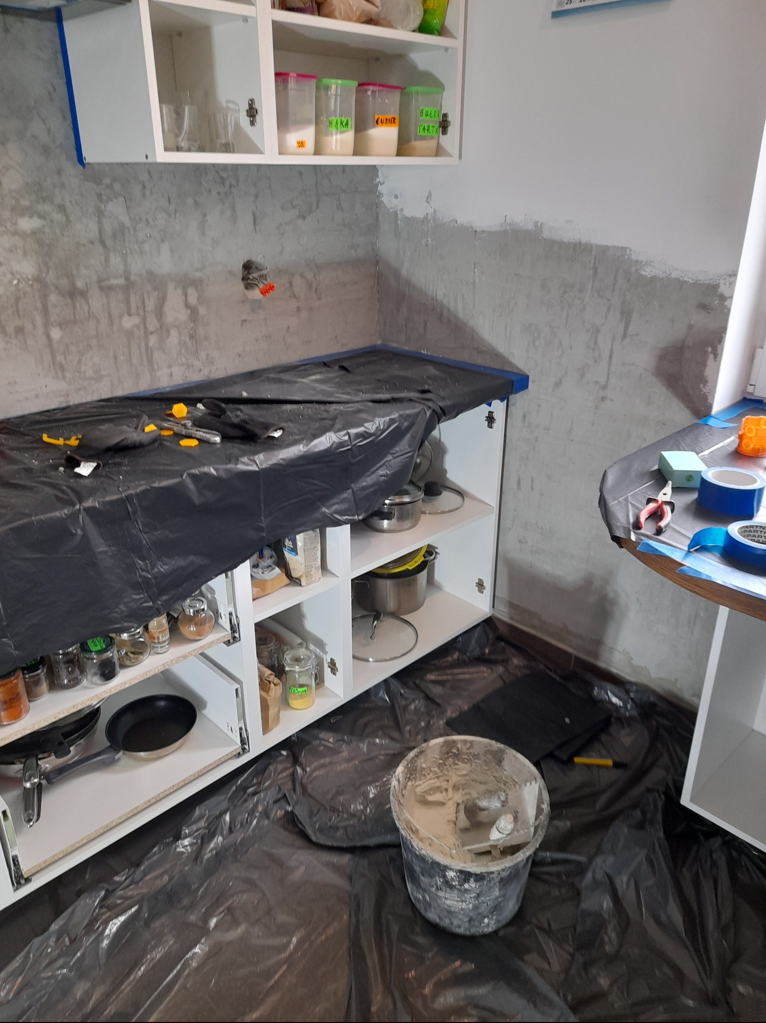 Przygotowanie kuchni do prac wykończeniowych: ściana w trakcie gruntowania, zabezpieczone folią meble kuchenne, wiadro z zaprawą i narzędzia na podłodze.