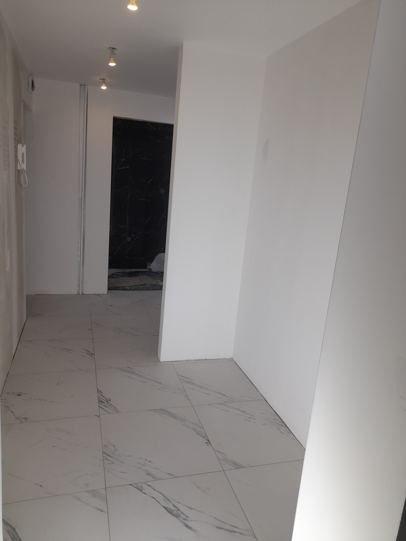 Korytarz w trakcie remontu: białe ściany, marmurowe płytki podłogowe, widok na łazienkę z czarnymi marmurowymi ścianami w głębi.