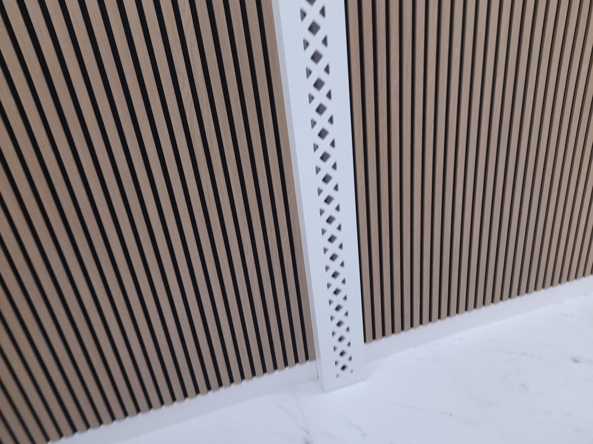 Ściana wykończona panelami drewnopodobnymi z czarnymi wstawkami, osłonięta białą, ażurową listwą narożną, na tle białej marmurowej podłogi.