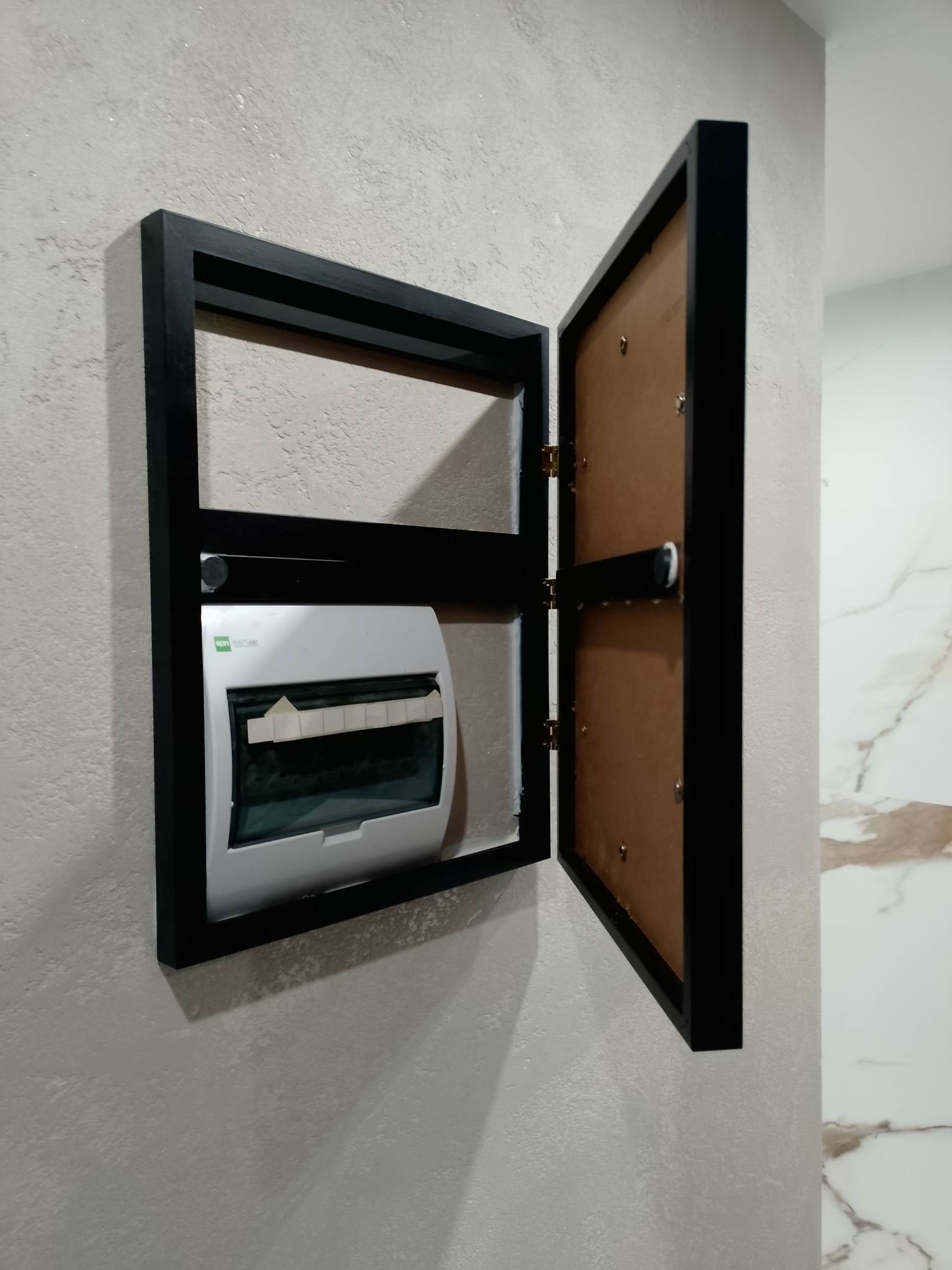 Otwarta, czarna ramka maskująca skrzynkę elektryczną na teksturowanej ścianie. Widoczna biała skrzynka z bezpiecznikami. Częściowo widoczna marmurowa ściana.