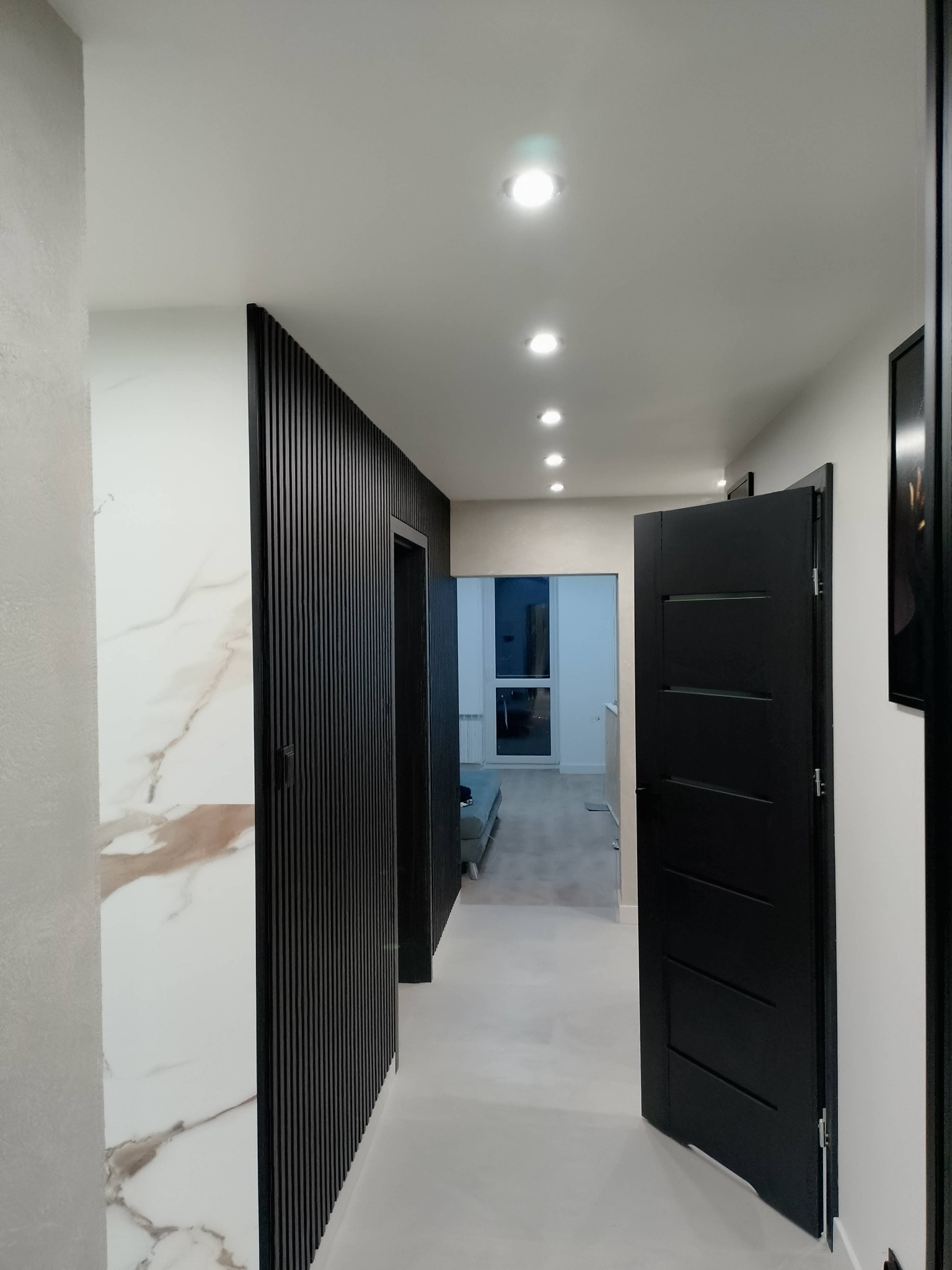 Nowoczesny korytarz po remoncie w Gostyninie: białe ściany z marmurowym akcentem, czarne lamele i drzwi, oświetlenie punktowe. Minimalistyczny design.