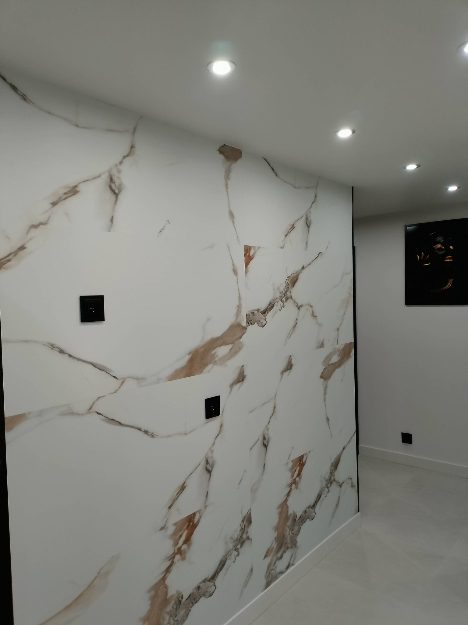 Ściana wykończona płytami imitującymi marmur z widocznymi gniazdkami elektrycznymi. W tle fragment korytarza z oświetleniem punktowym i obrazem na ścianie.