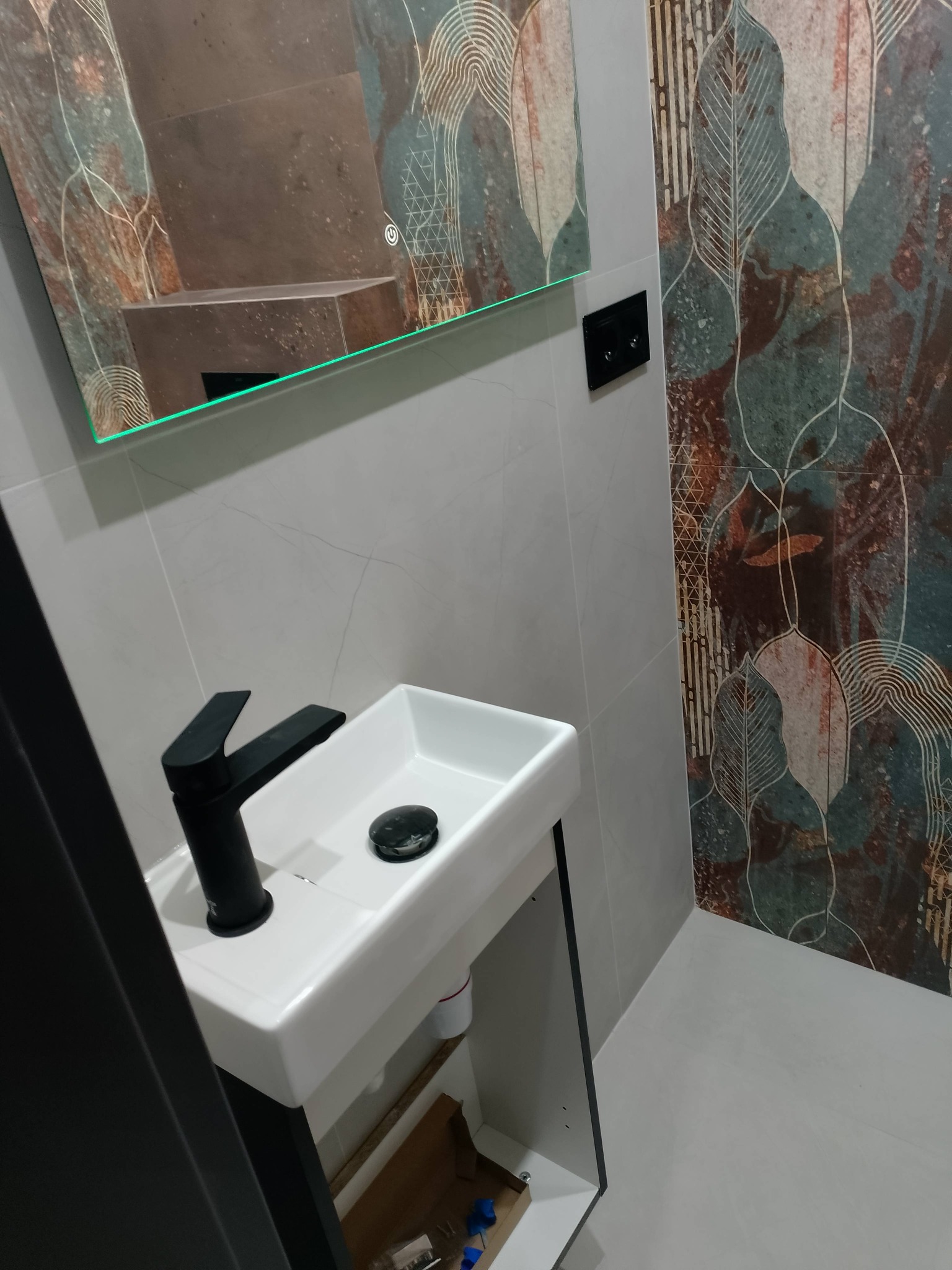 Nowoczesna łazienka: biała umywalka z czarną armaturą, lustro z podświetleniem i dekoracyjne płytki ścienne z motywem liści w odcieniach brązu i zieleni.